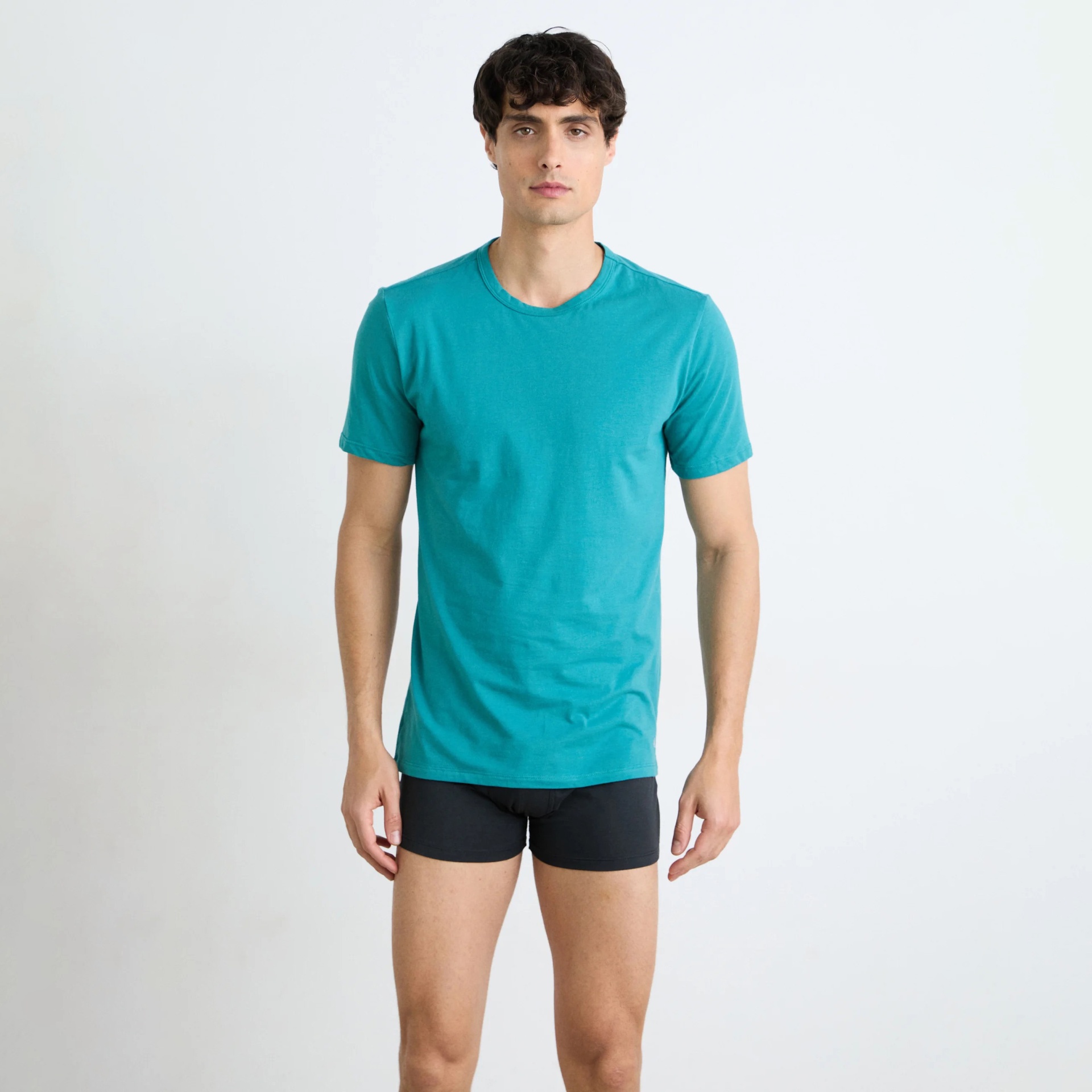 Homem com t-shirt azul turquesa de manga curta e cuecas boxer pretas em fundo branco