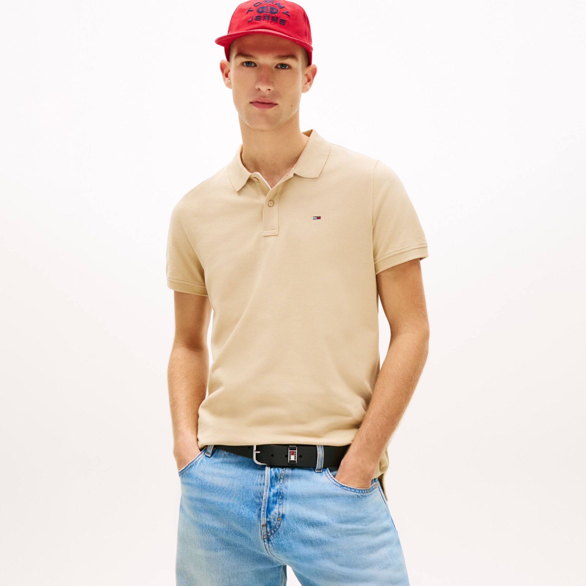 Modelo a usar polo bege, calças de ganga azuis claras, cinto preto e boné vermelho com texto