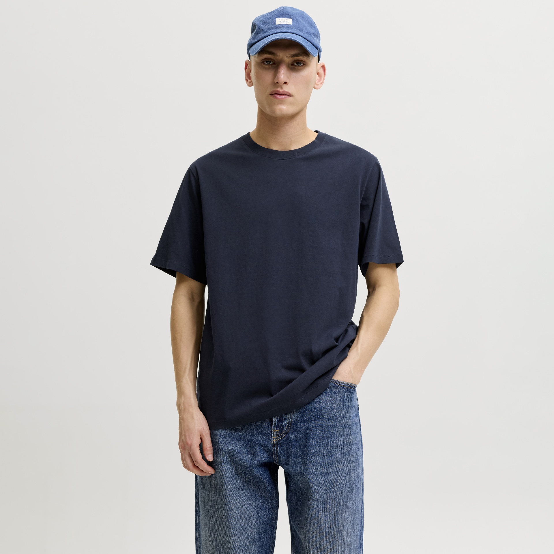 Homem com t-shirt azul escura, calças de ganga azuis e boné azul com etiqueta JACK & JONES