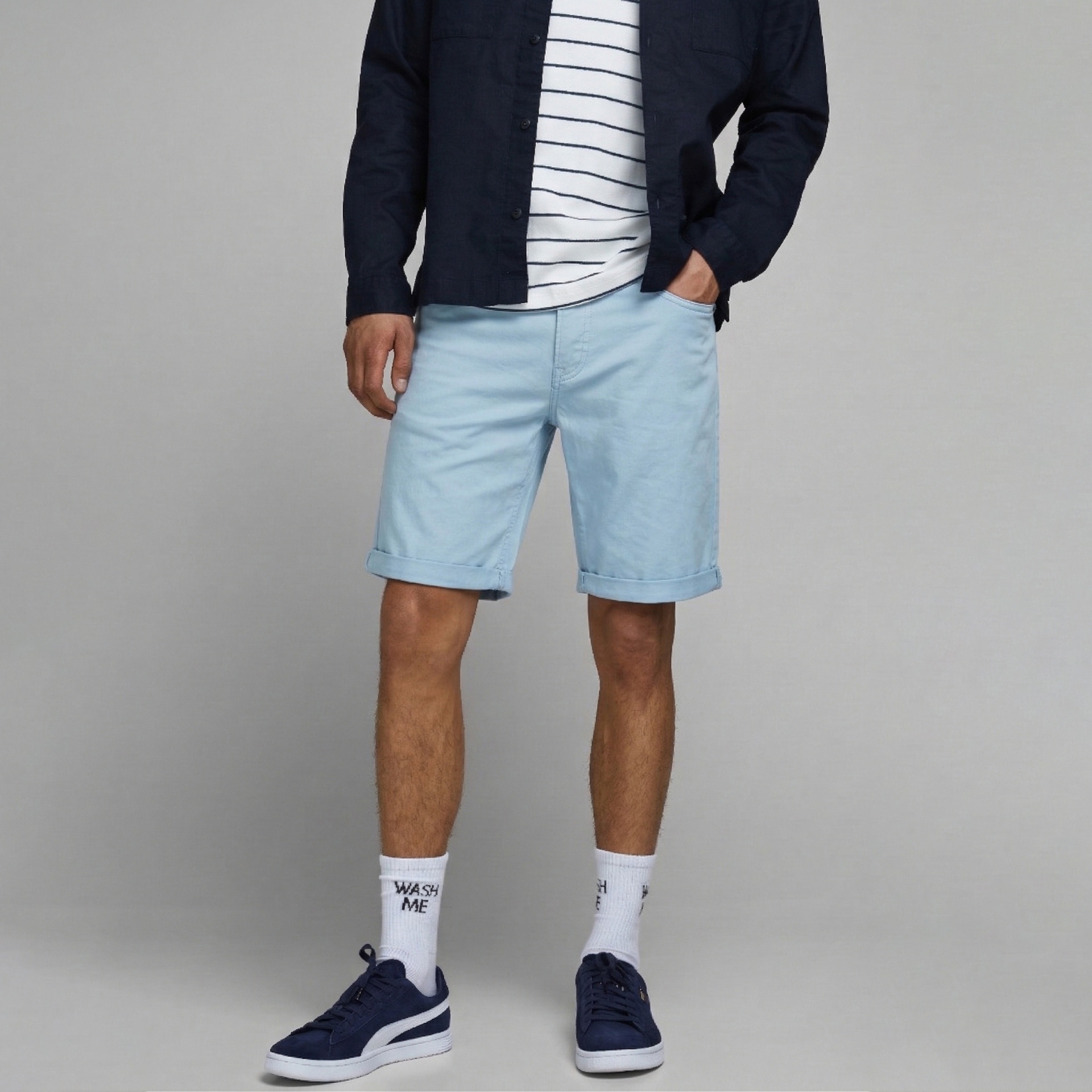 Calções azuis claros com bainha virada usados por homem com camisa azul escuro e t-shirt às riscas, meias com texto e sapatilhas azuis.