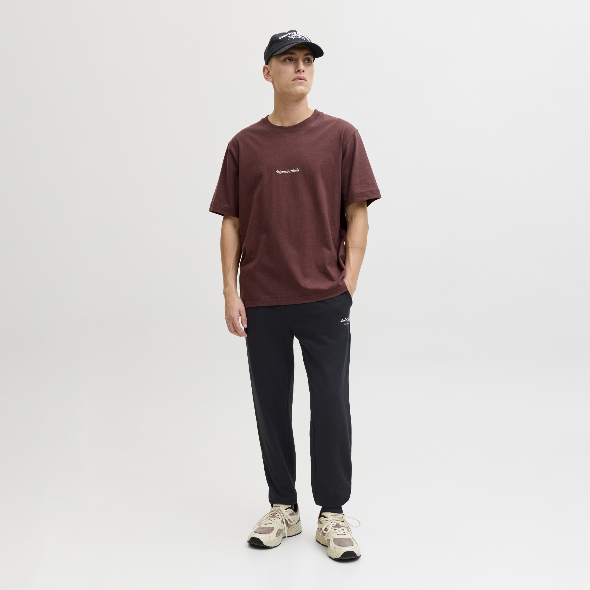 Homem com t-shirt castanha, calças pretas, boné preto e sapatos bege