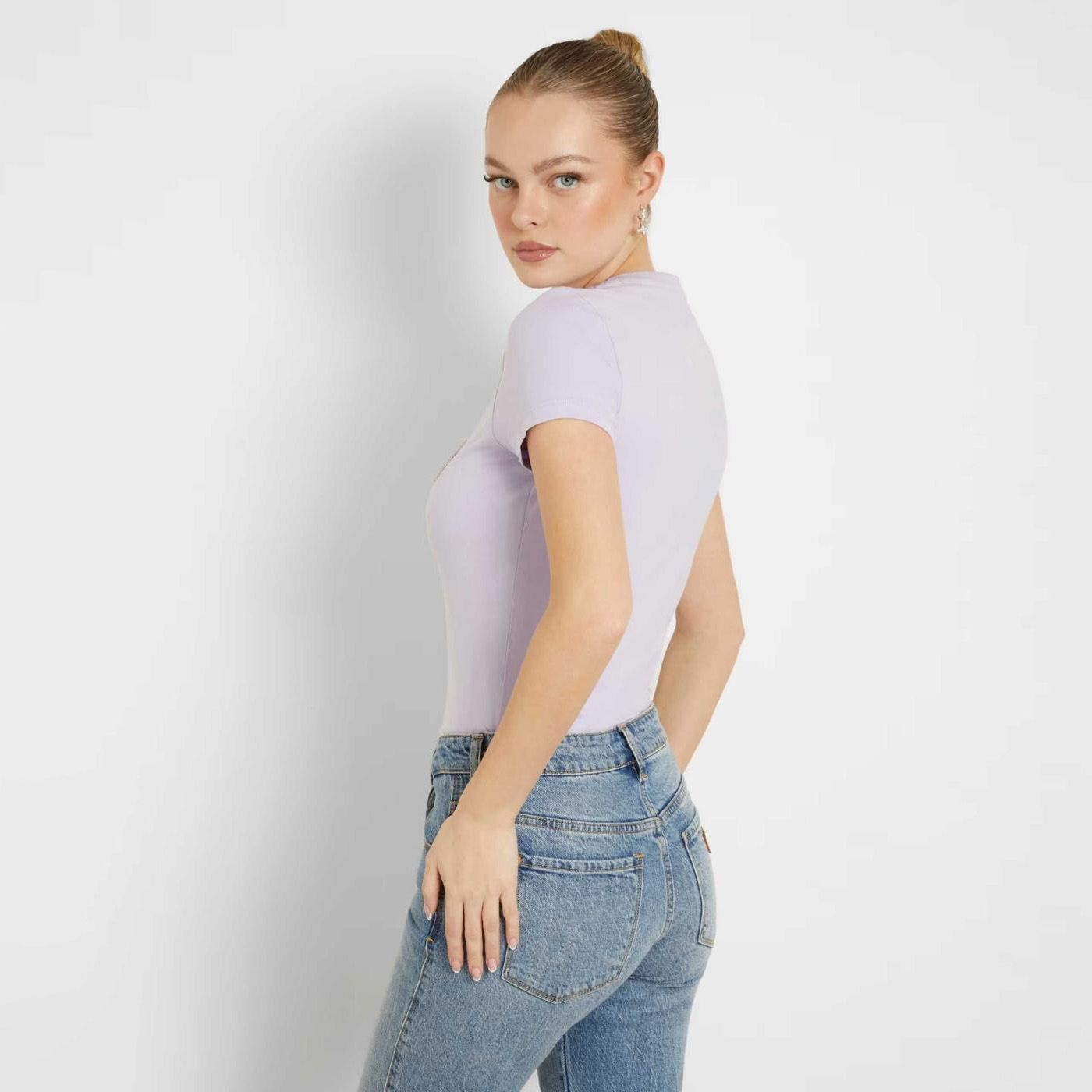 Mulher de perfil com camiseta lilás e jeans azul claro em fundo branco