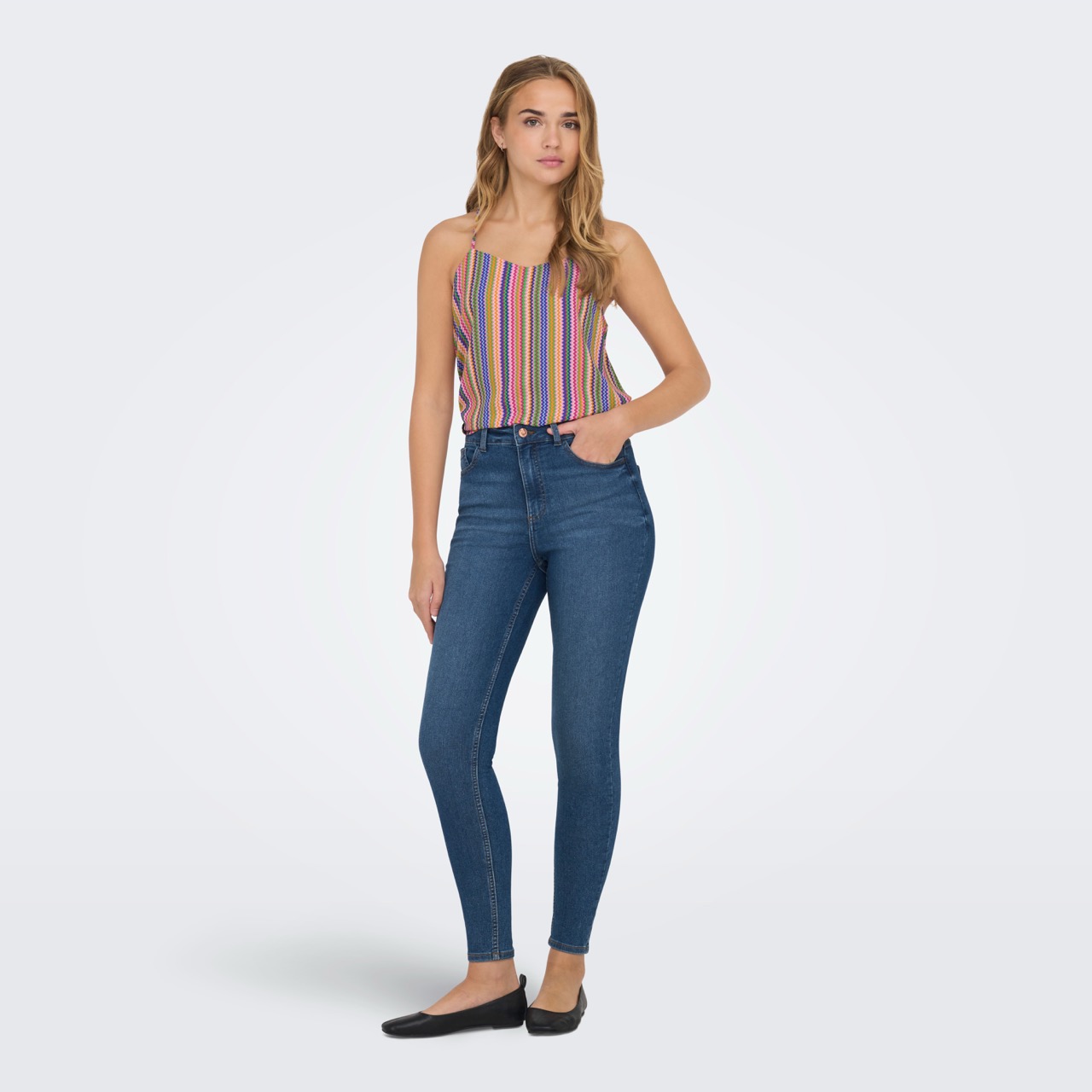 Modelo feminina veste calças de ganga azul escura skinny e top colorido às riscas verticais.