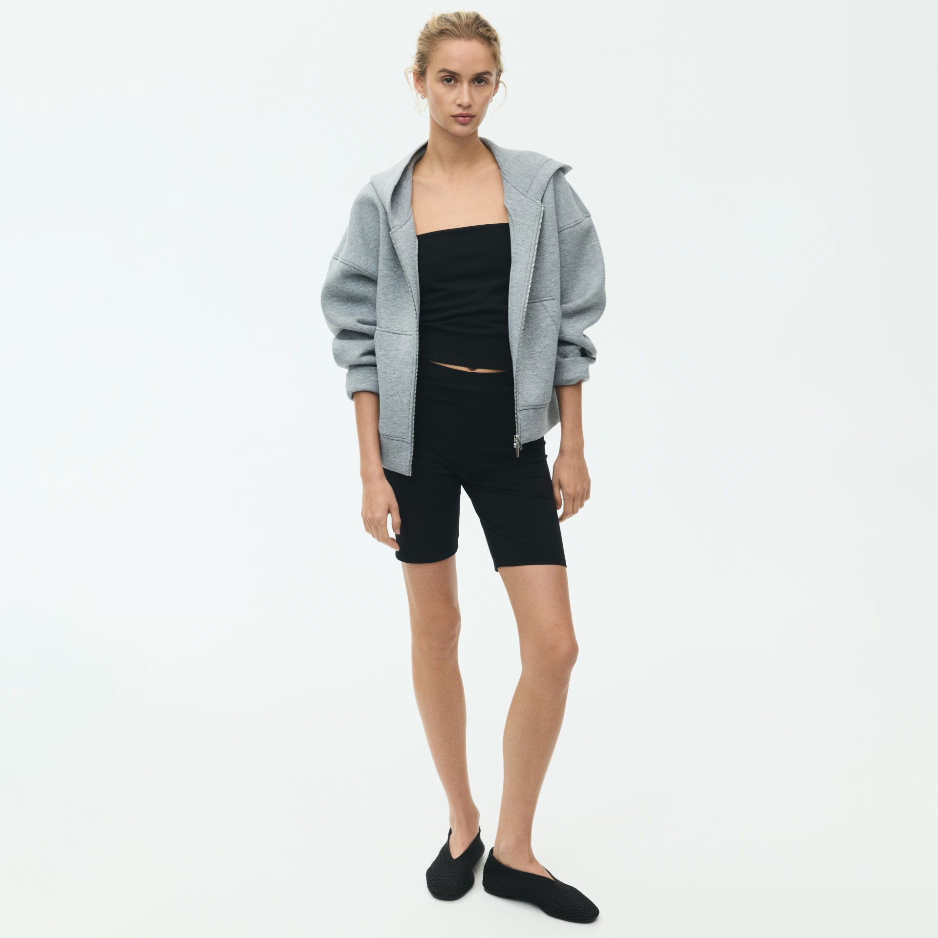 Mulher com casaco cinza, top preto, calções pretos e sapatos pretos em fundo branco
