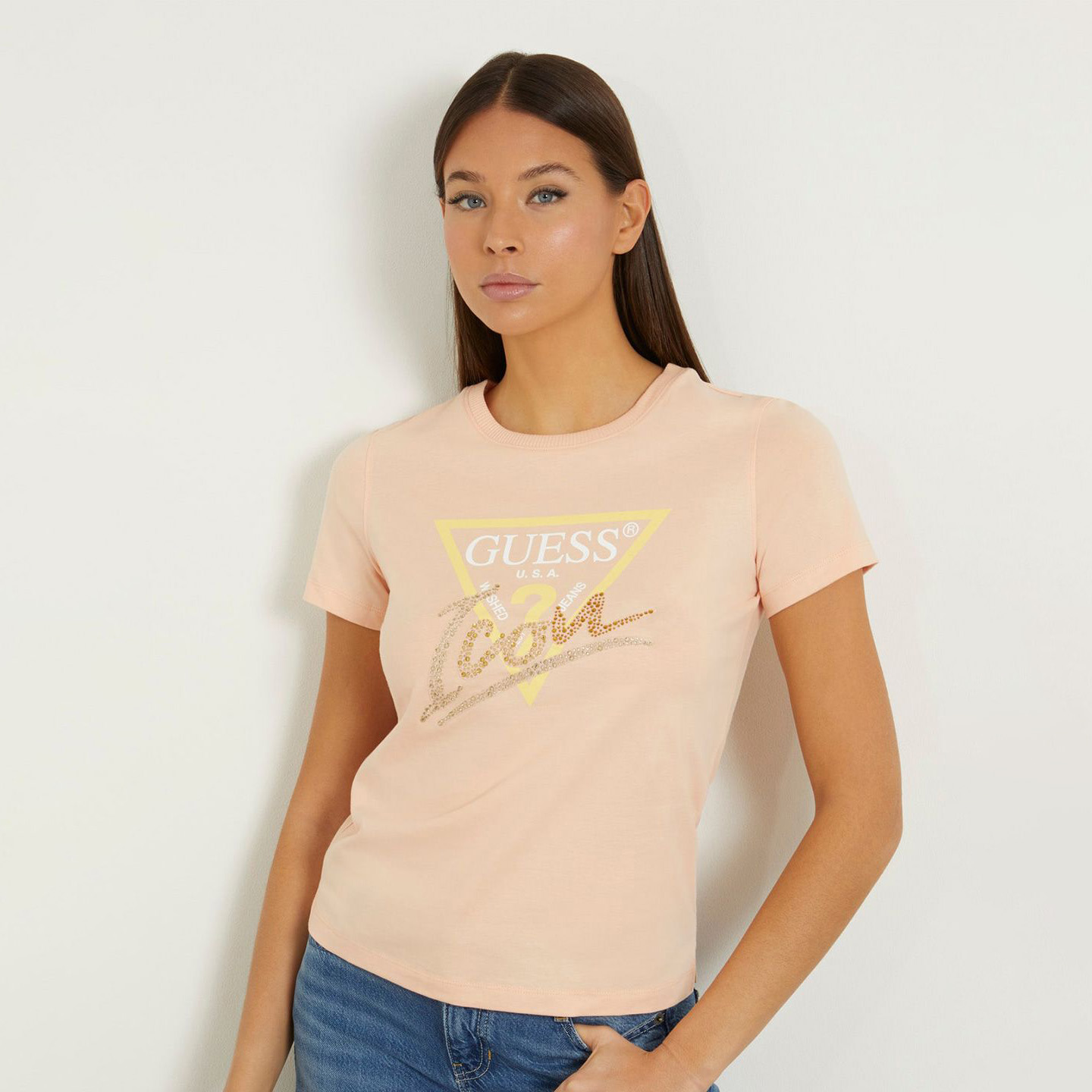 T-shirt rosa claro com logo GUESS e palavra Icon brilhante, usada por mulher com jeans azuis