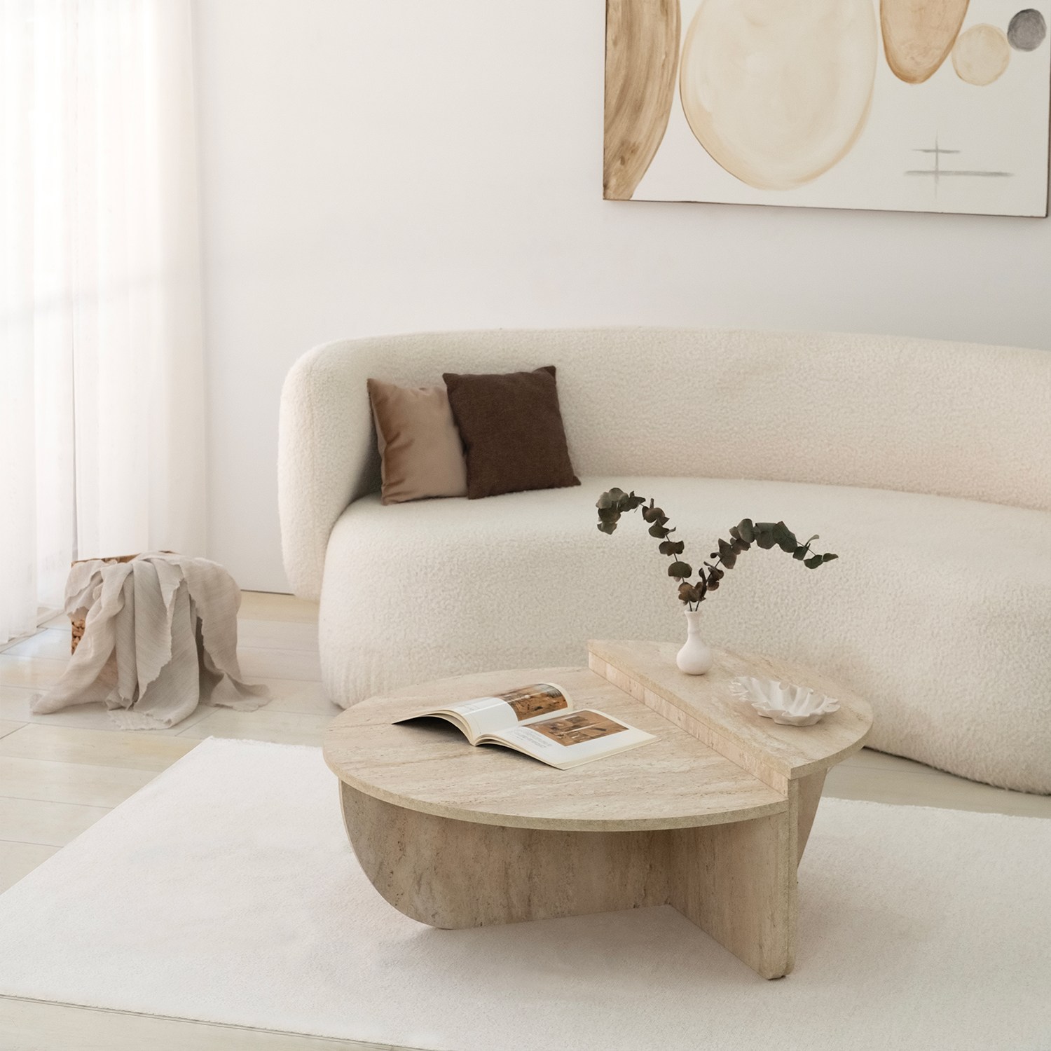 Mesa de centro em madeira clara com vaso branco e folhas secas, sofá branco curvo e almofadas castanhas.