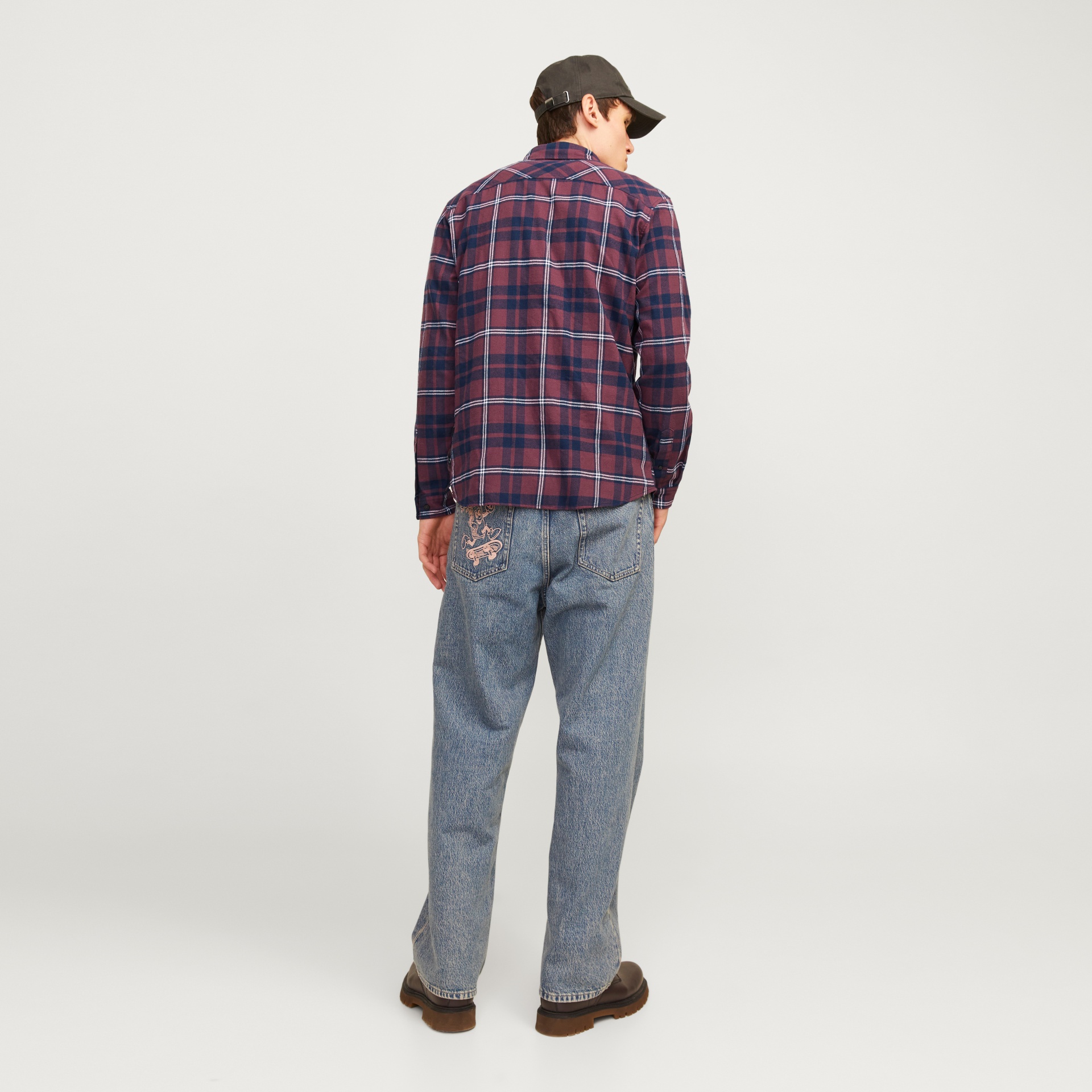 Homem de costas com camisa xadrez, calças de ganga bordadas e boné preto.