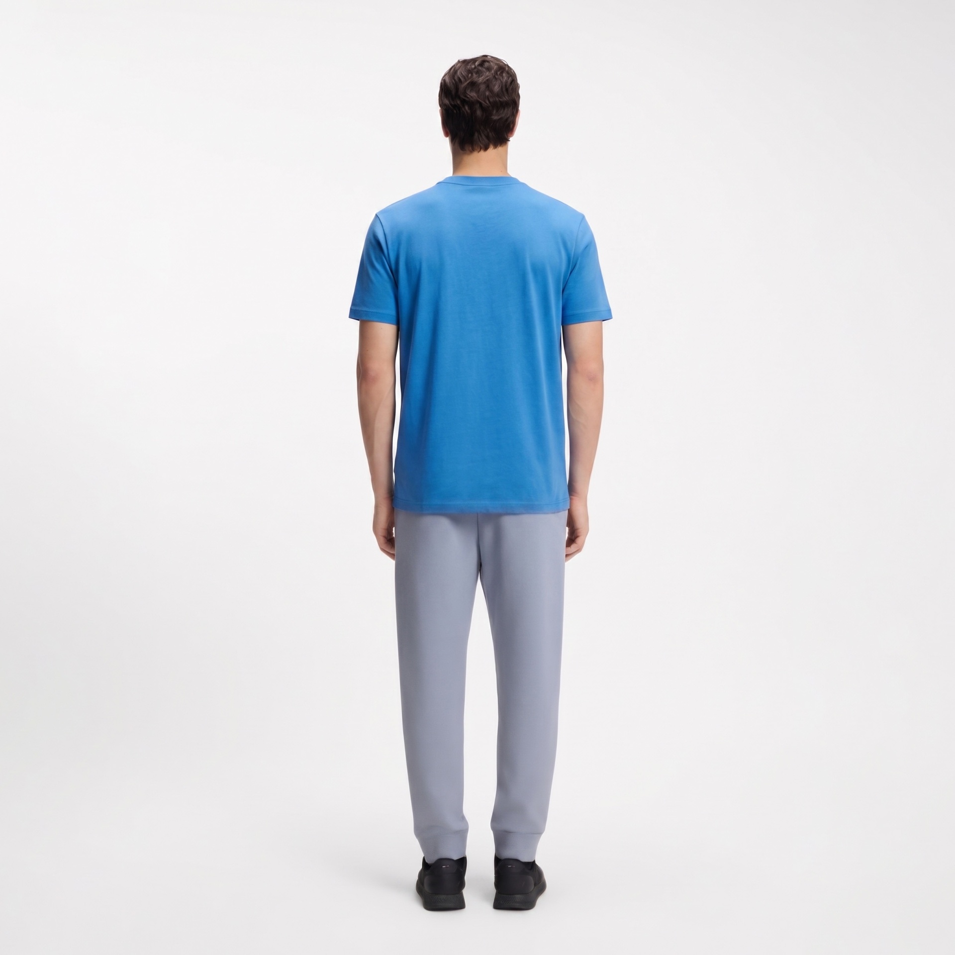 Pessoa de costas com t-shirt azul, calças cinza e sapatos pretos