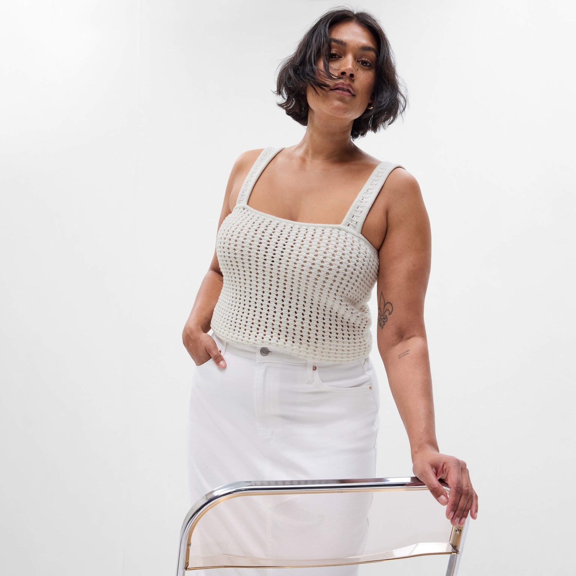 Mulher a usar top branco de malha com alças largas e saia branca em estúdio com fundo branco e cadeira transparente