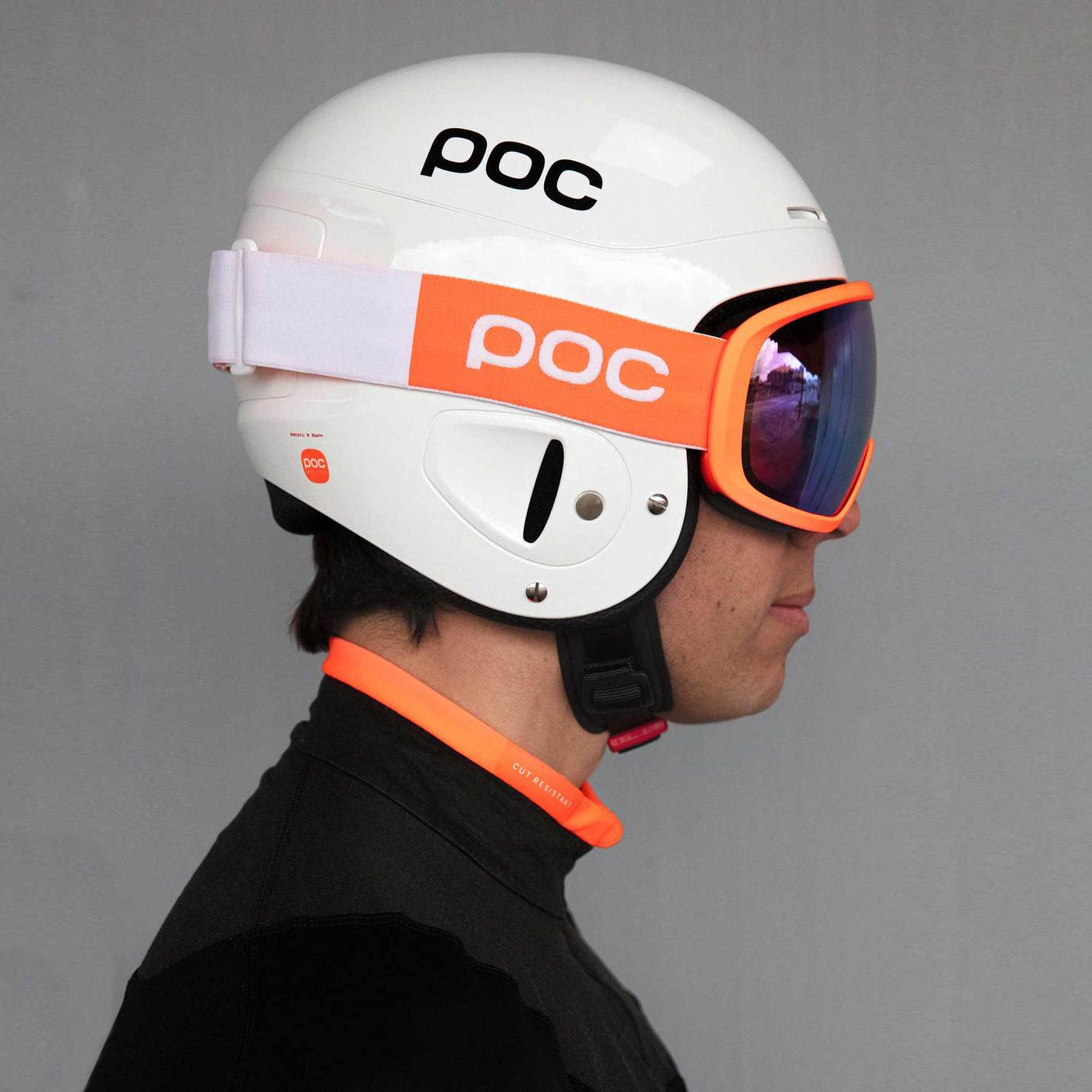 Capacete branco com óculos de proteção laranja e azul e faixa elástica com logo 'poc', usado por pessoa com camisa preta de gola laranja.