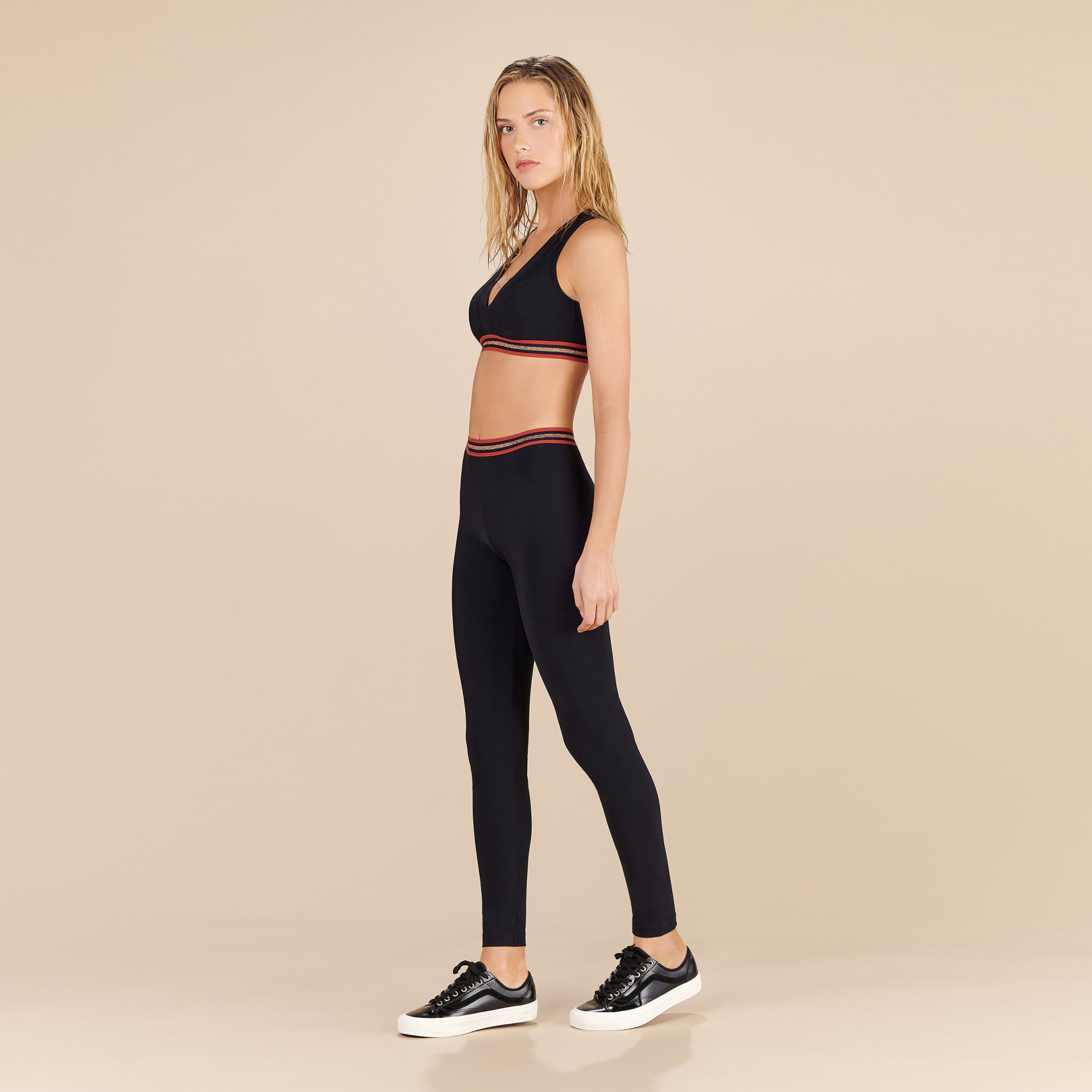 Mulher com conjunto desportivo preto com faixas vermelhas e brancas, sapatilhas pretas, fundo bege