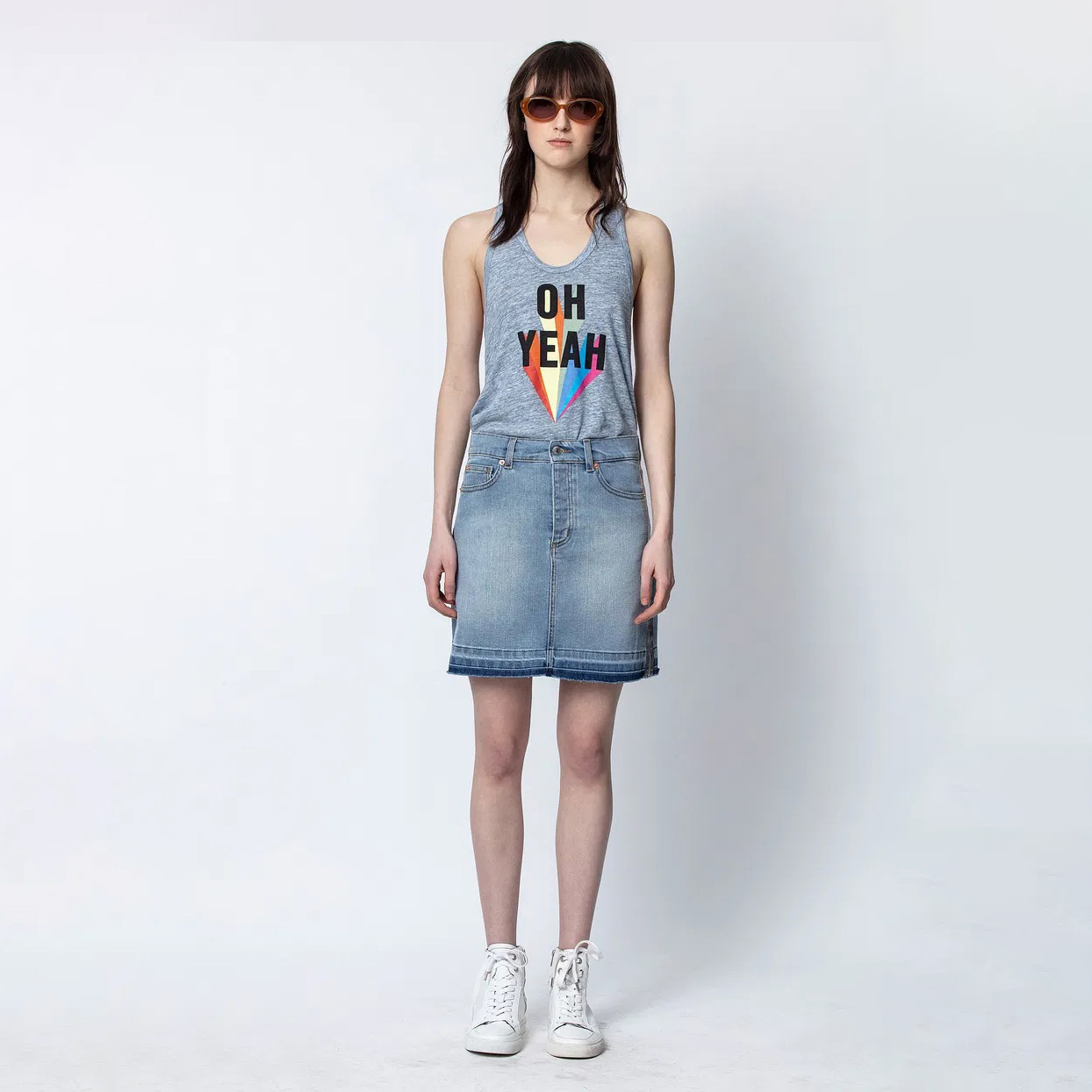 Mulher com camisola cinzenta estampada, saia de ganga azul e tênis brancos em fundo branco.
