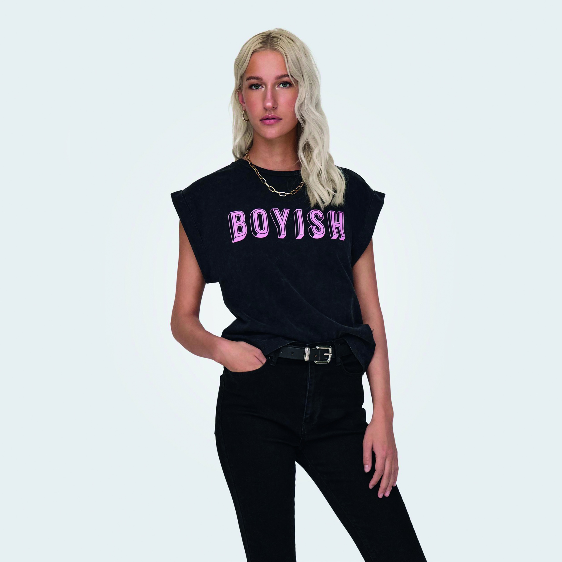 Blusa preta com texto BOYISH em rosa, usada por mulher loira, calças pretas e fundo cinza