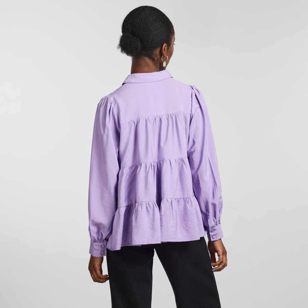 Blusa lilás de mulher vista de costas com babados, mangas compridas e punhos abotoados.