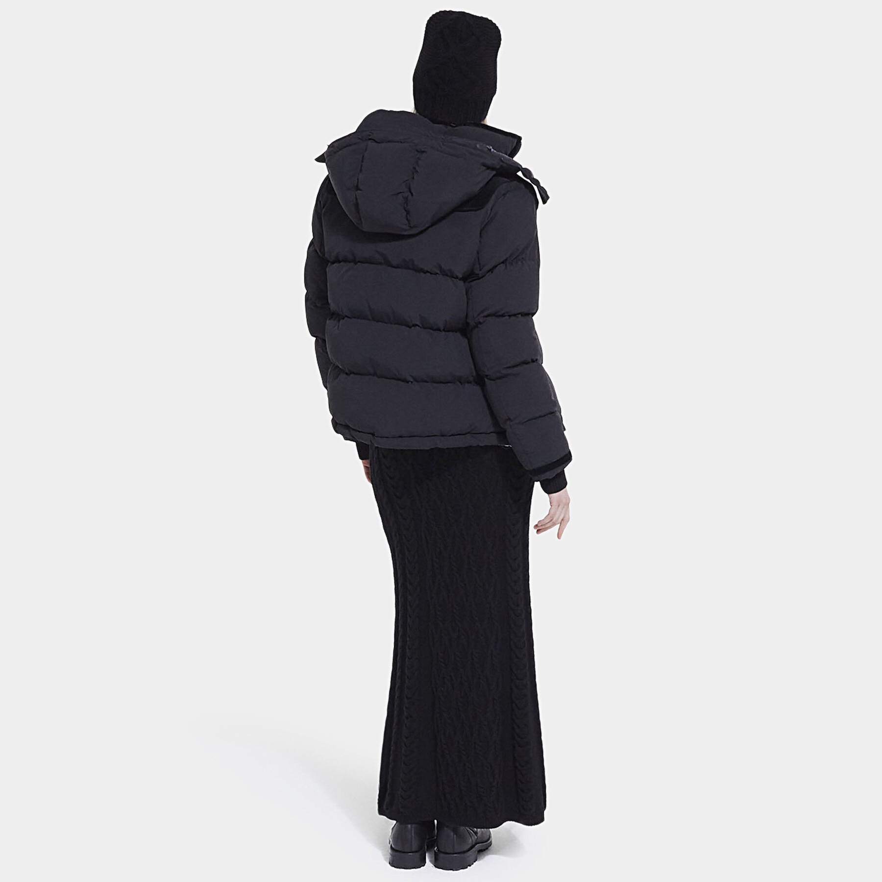 Pessoa de costas com casaco preto acolchoado, saia de malha longa preta, botas e gorro preto.