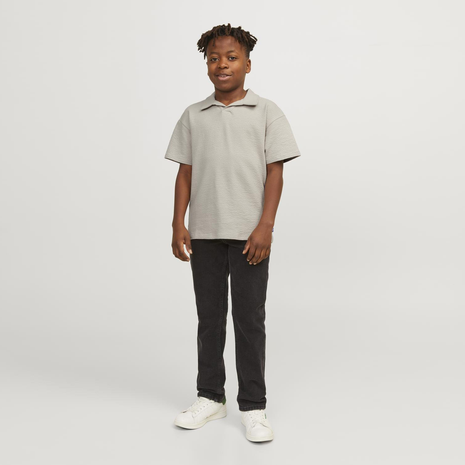 Rapaz com camisa cinza claro e calças pretas posando em fundo branco.