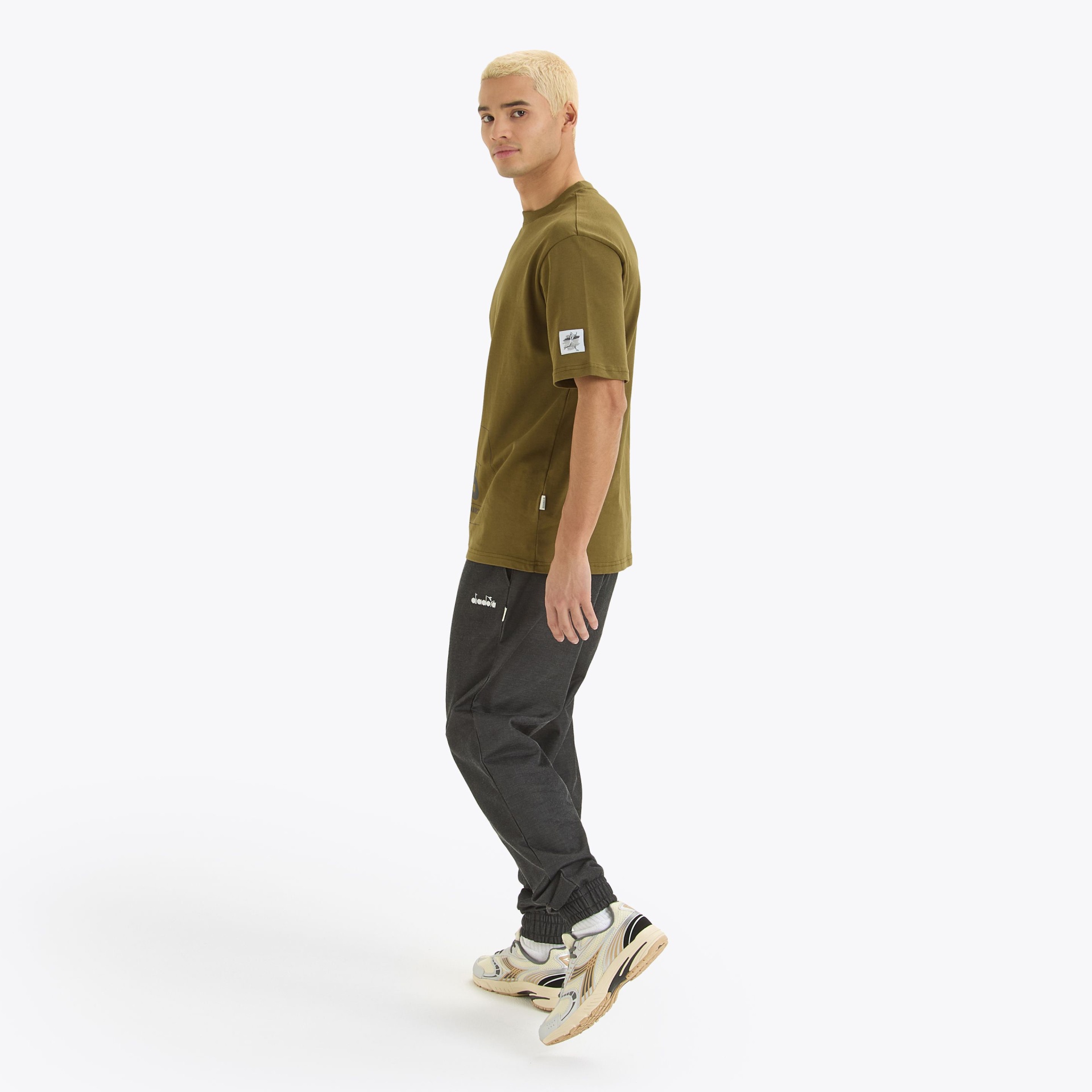 Homem de perfil com t-shirt verde-oliva, calças negras e ténis branco e bege.