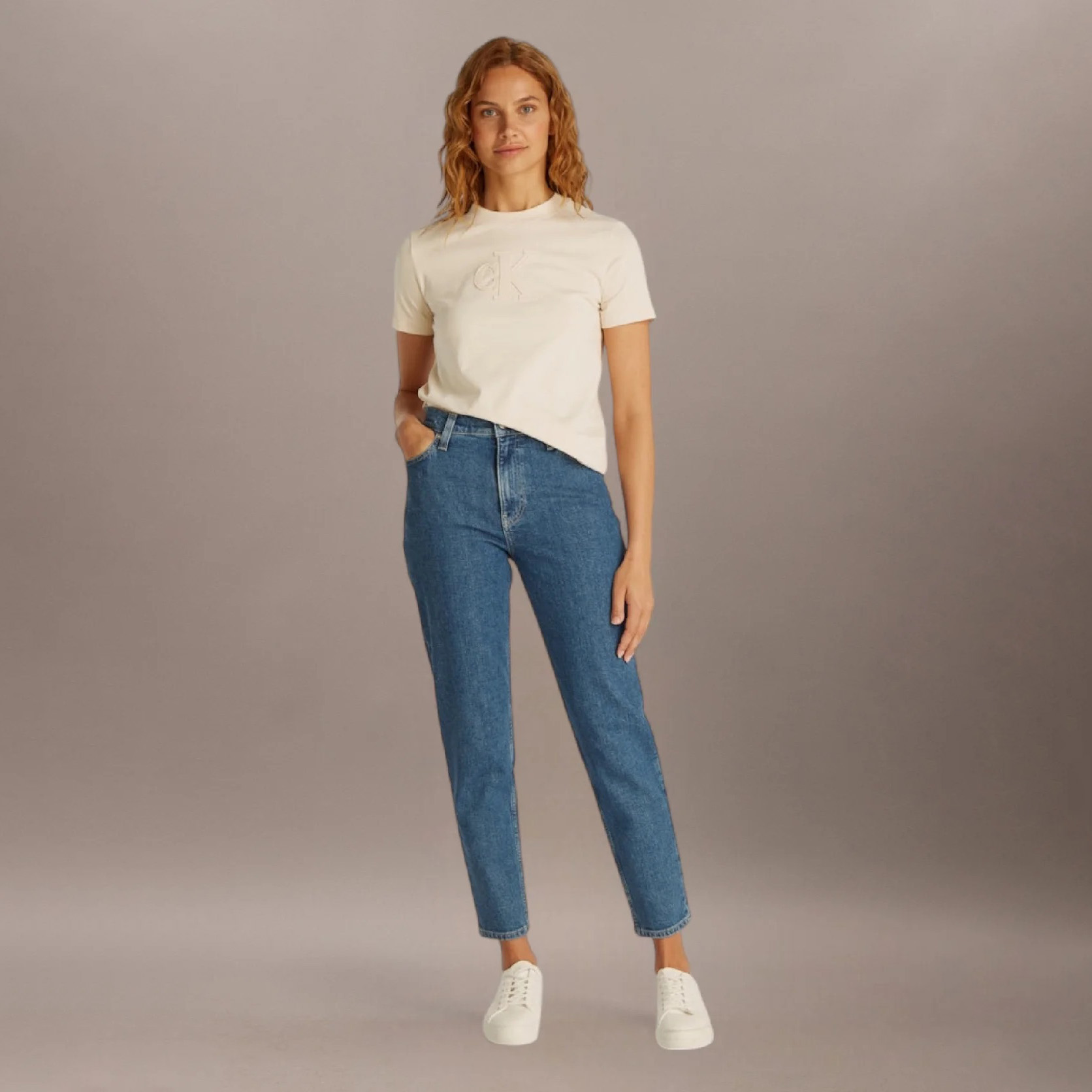 Mulher veste t-shirt branca com logotipo ck em relevo e jeans azul escuro, sapatos brancos, fundo cinza.