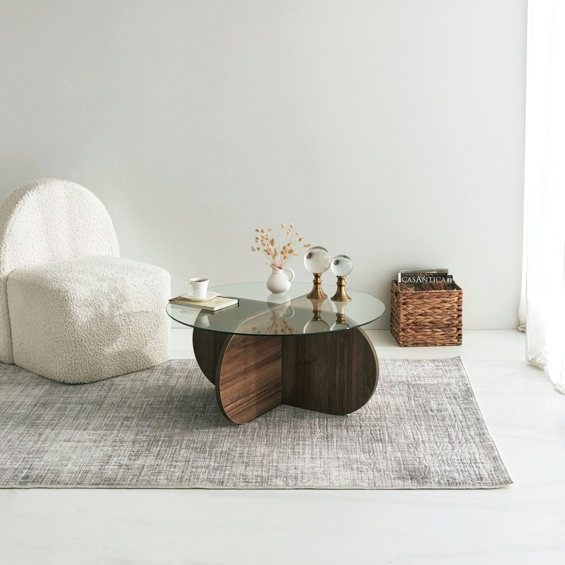 Conjunto de sala com cadeira branca texturizada, mesa de vidro com base em madeira, acessórios decorativos e tapete cinza em ambiente claro