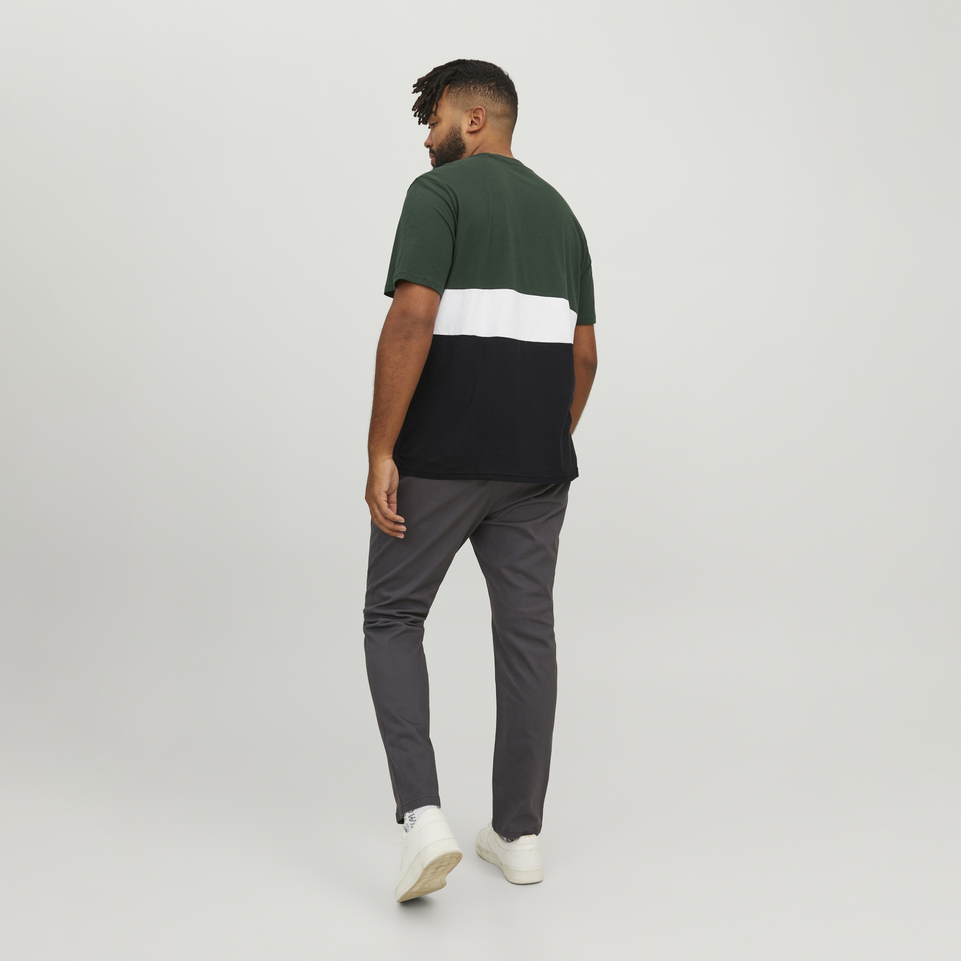 Homem de costas com t-shirt bicolor verde, branco e preto, calças cinza e sapatilhas brancas.