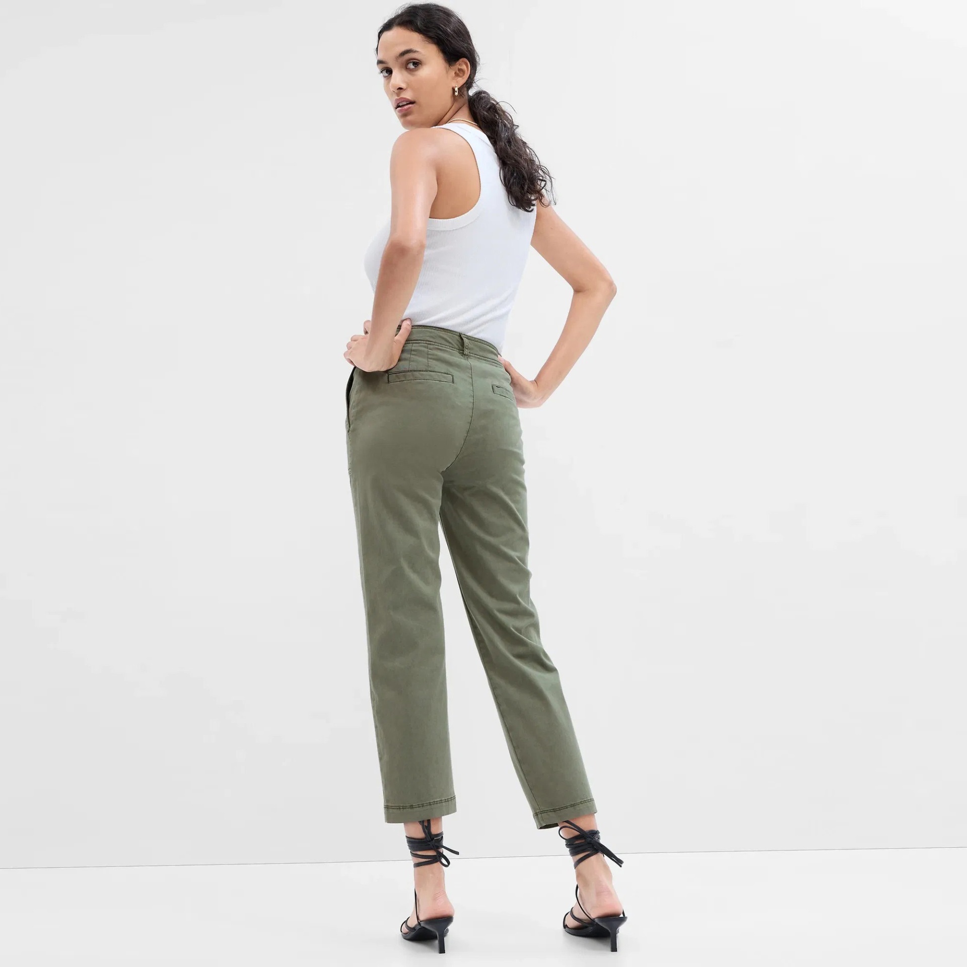 Mulher com calças verdes de corte reto, t-shirt branca e sandálias pretas em fundo branco