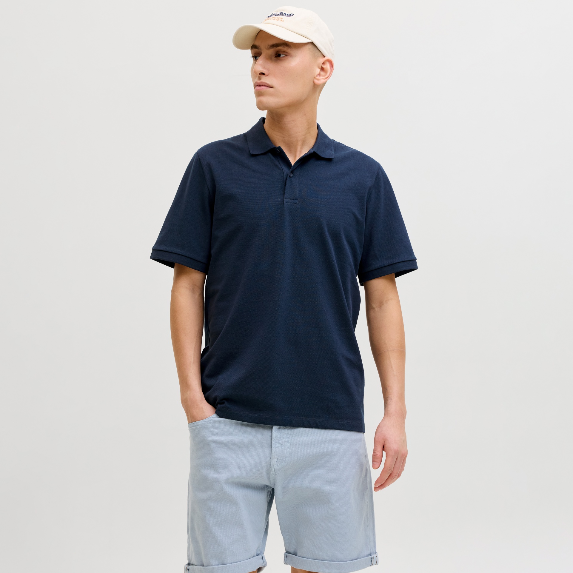 Homem com polo azul-marinho, calções azul-claro e boné bege com texto