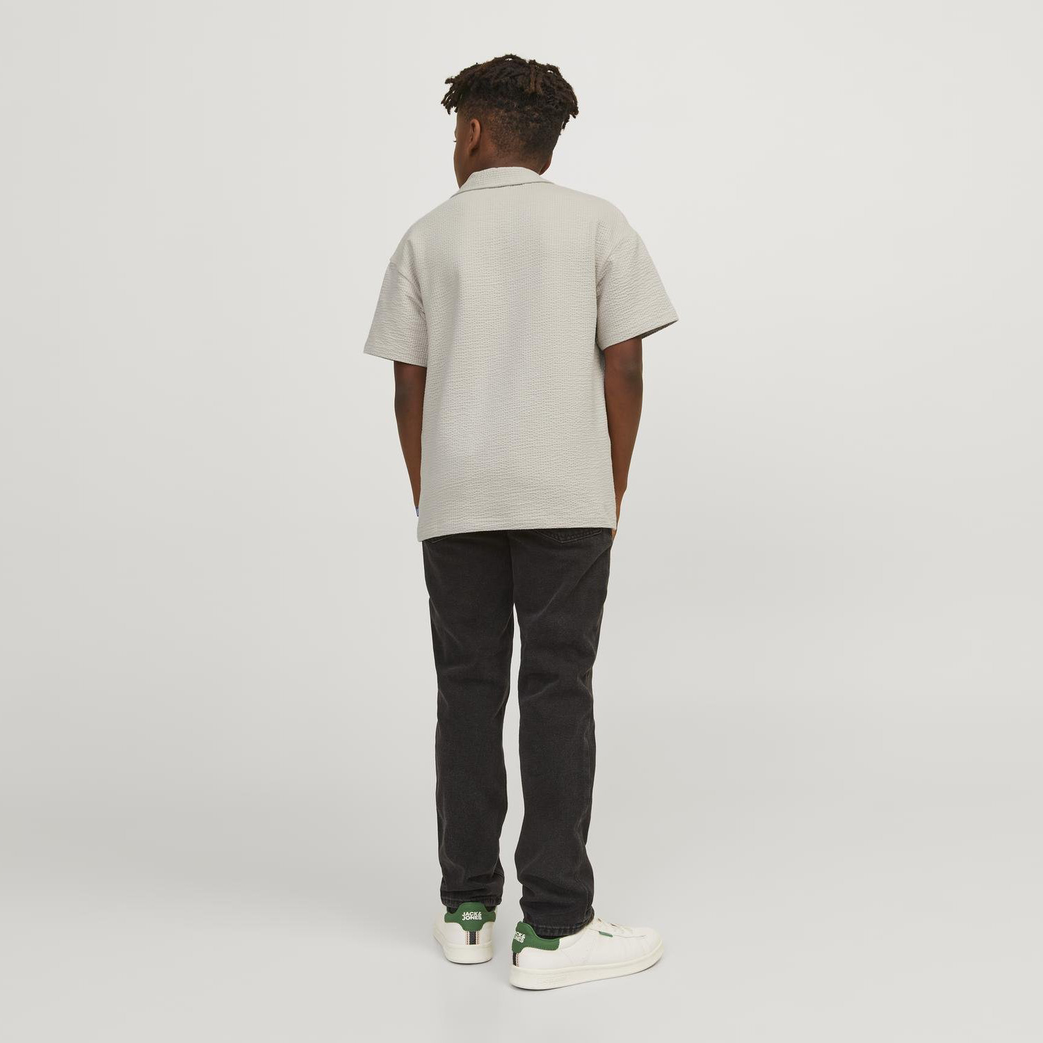 Homem de costas com camisa bege, calças pretas e ténis brancos com detalhes verdes