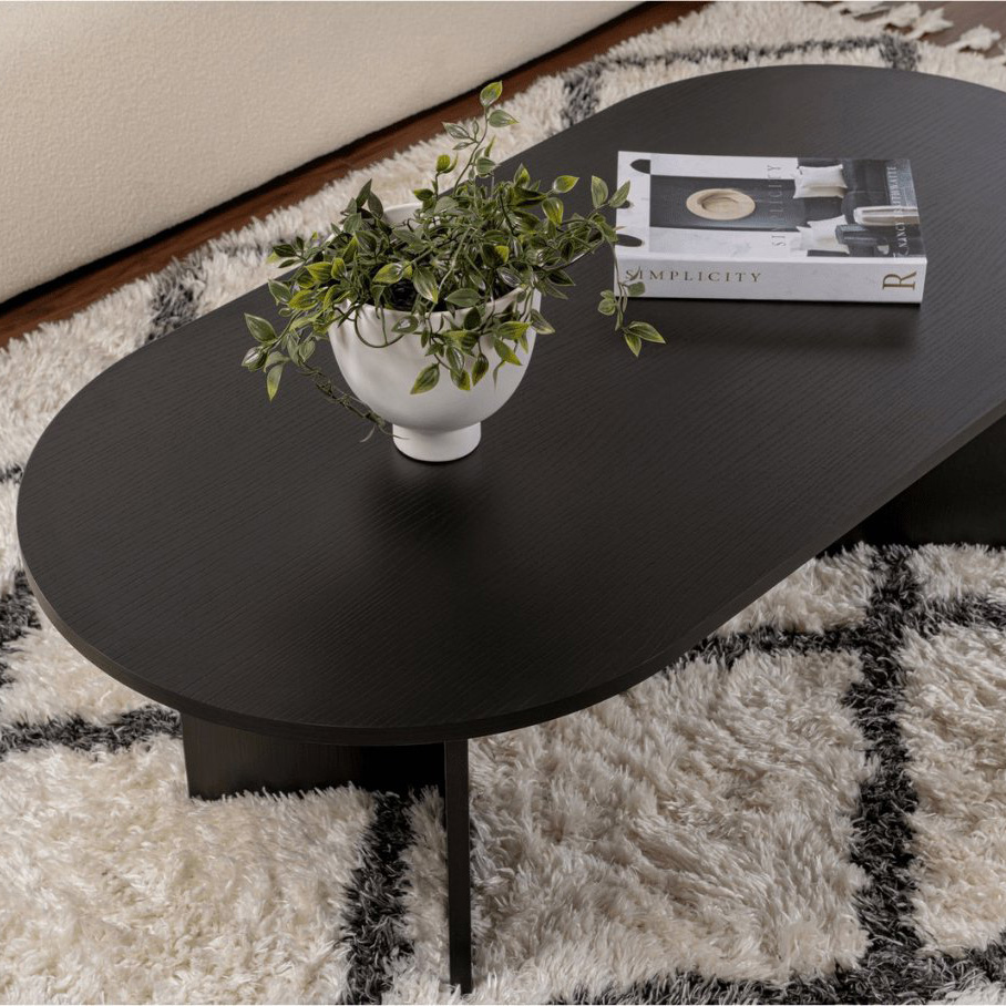 Mesa de centro preta oval com planta e livro em cima, sobre tapete branco com padrão xadrez preto