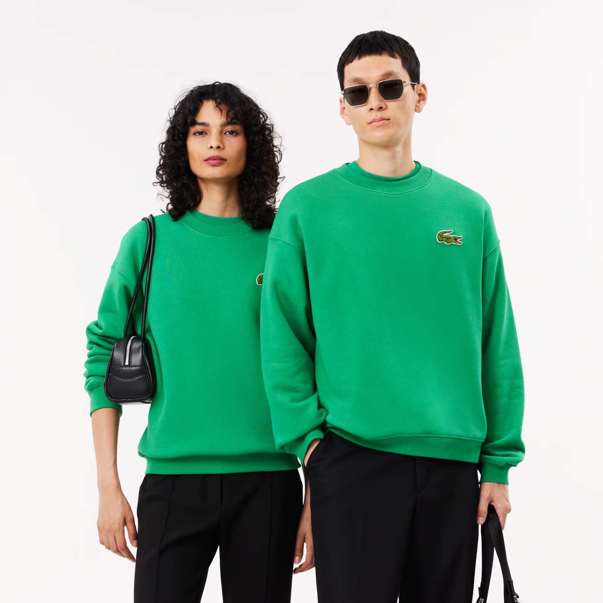Duas pessoas com sweatshirts verdes Lacoste, calças pretas, mulher com mala preta e homem com óculos escuros e mala.