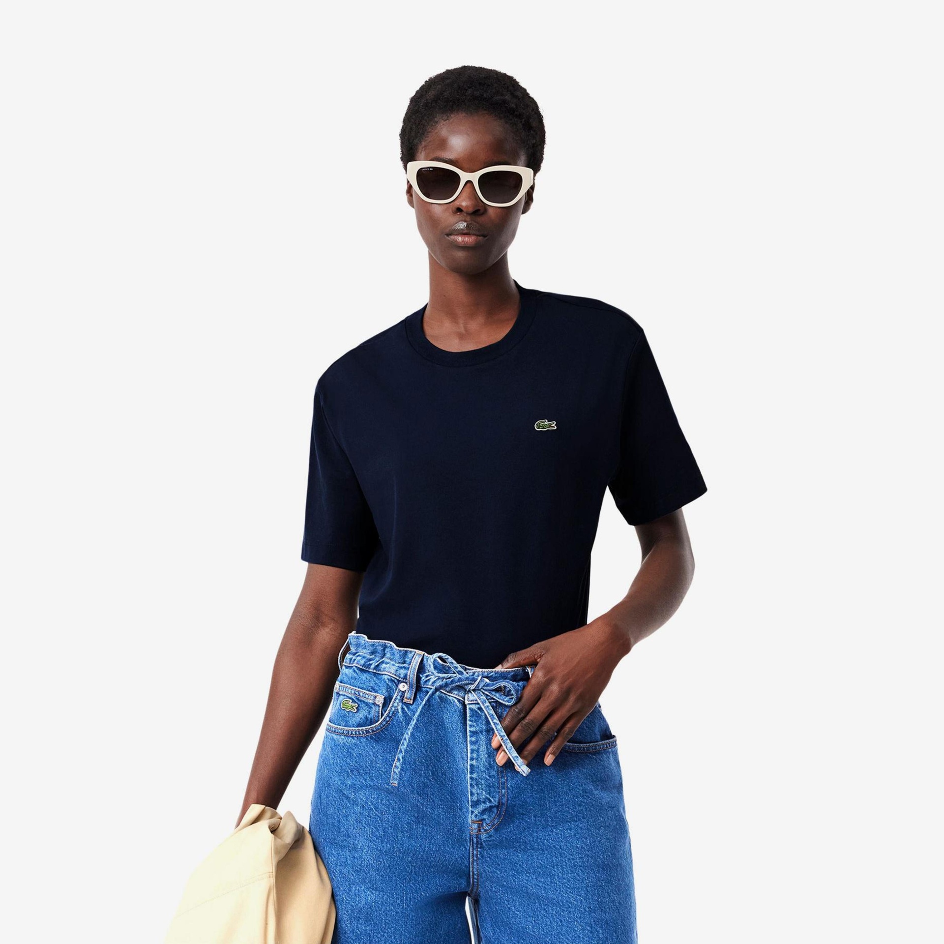 Pessoa com t-shirt preta, calças de ganga azuis, óculos de sol brancos e roupa bege na mão