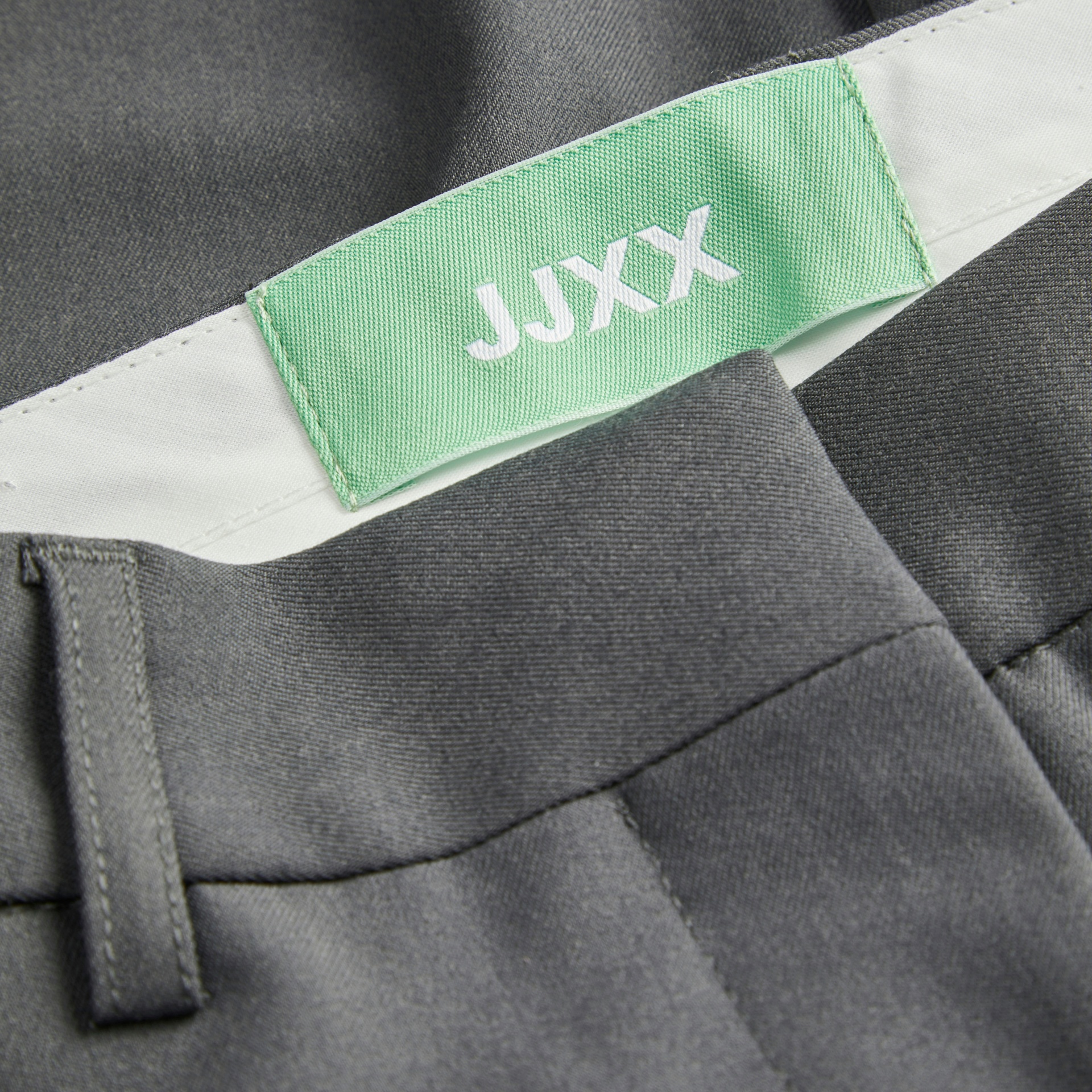 Etiqueta verde com texto JJXX numa peça de roupa cinzenta com cinto branco