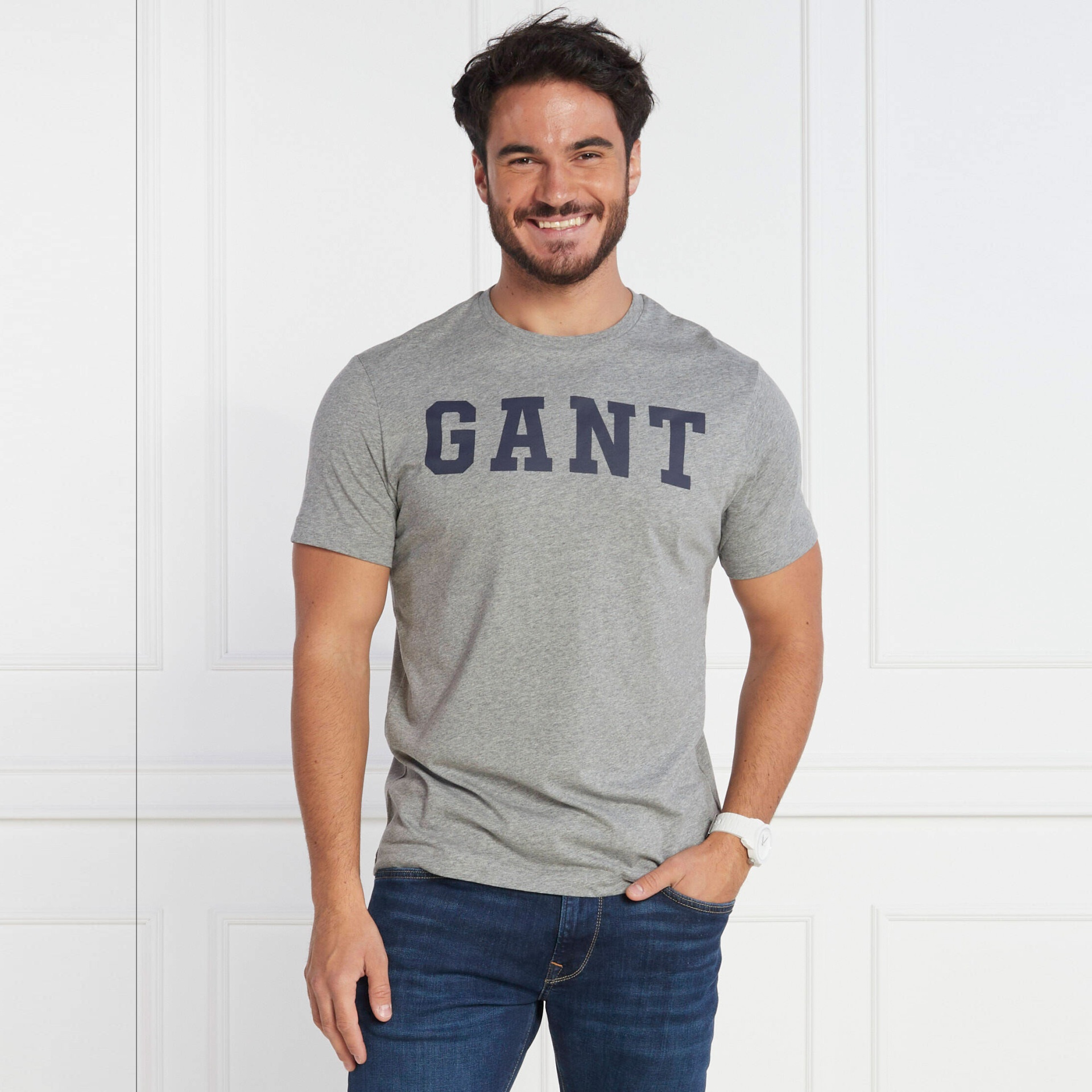 Homem a sorrir a usar t-shirt cinzenta com texto azul GANT e calças de ganga azuis num ambiente interior branco