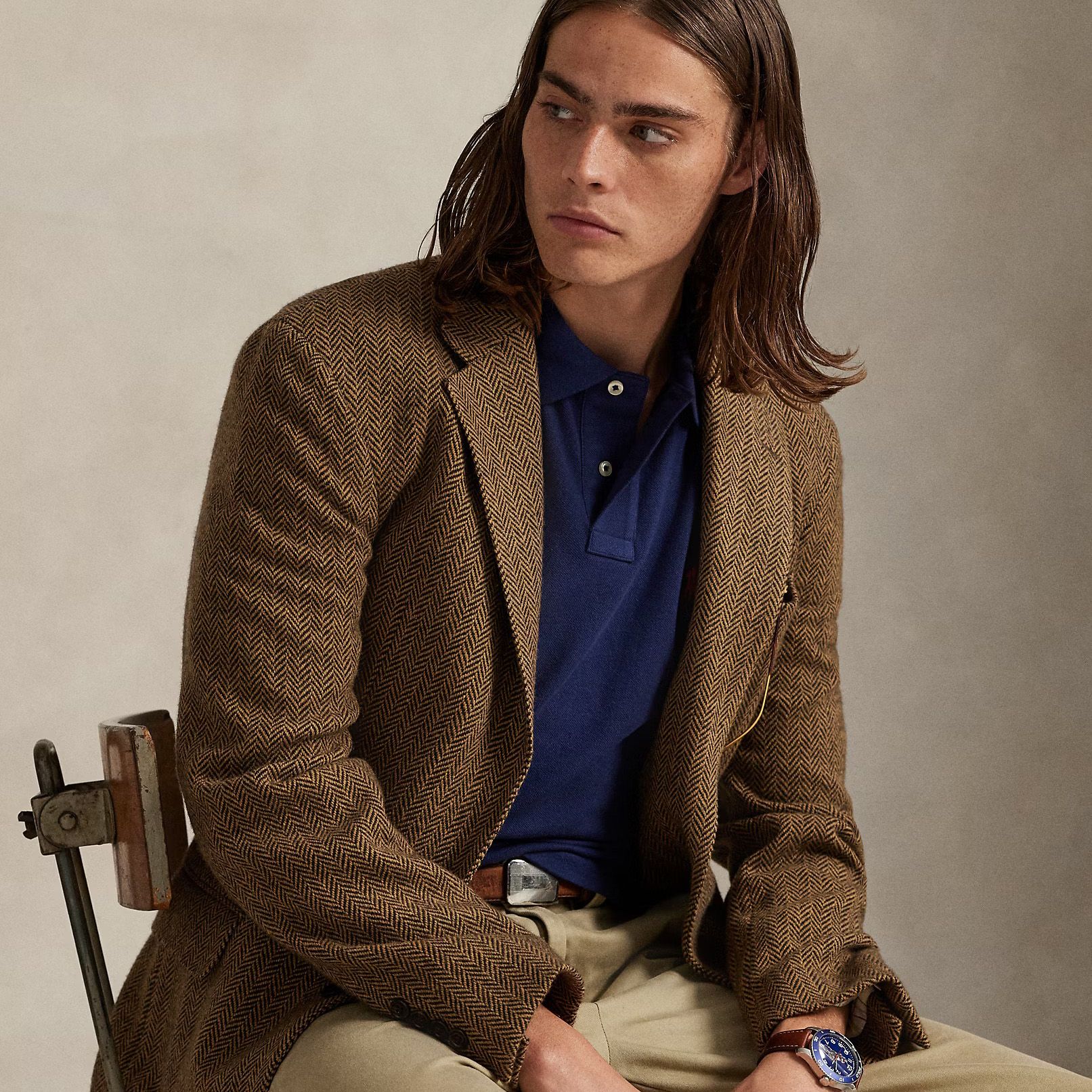 Homem vestido com blazer castanho padrão espinha de peixe, polo azul e calças bege sentado numa cadeira