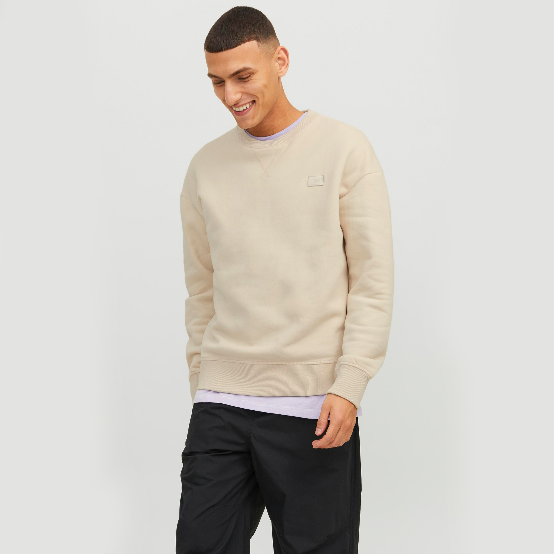 Homem sorridente usando sweatshirt bege com calças pretas em fundo cinza claro