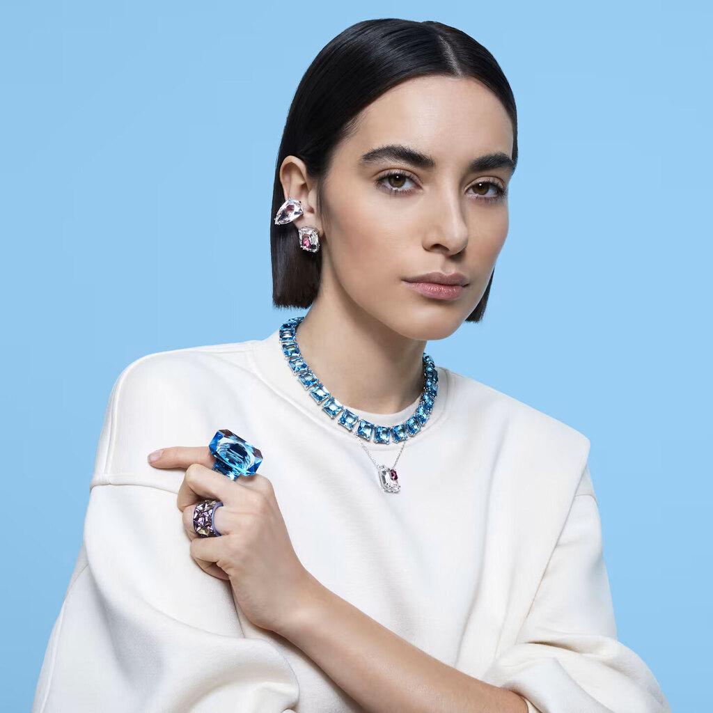 Mulher com colar, brincos e anéis de pedras coloridas, e camisola branca, com fundo azul.