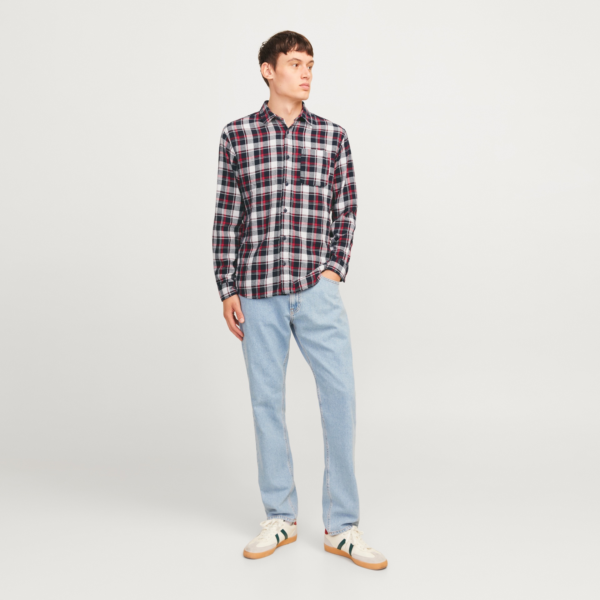 Homem com camisa xadrez azul, branca e vermelha, calças de ganga azul clara e sapatilhas brancas com detalhes verdes.