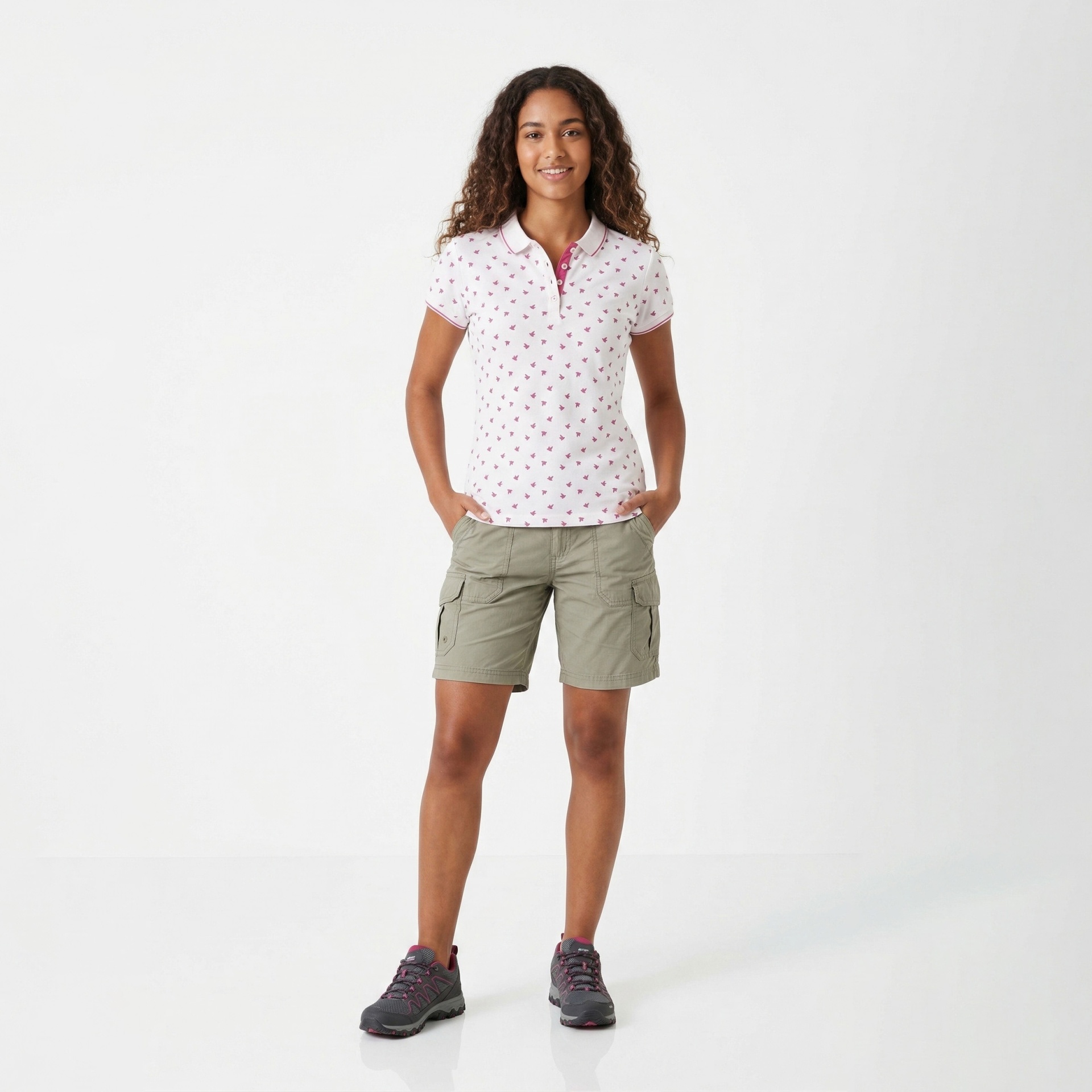 Mulher com polo branca com padrões rosa, calções verdes, sapatos cinzentos, fundo branco.