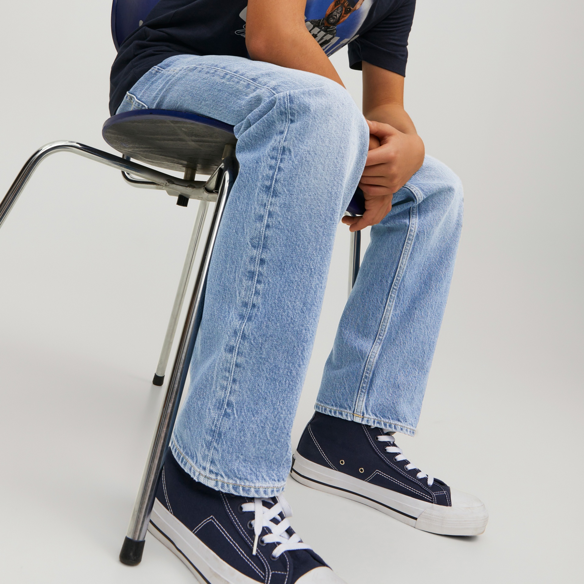 Pessoa sentada com calças de ganga azul claras e ténis de lona azul escura em cadeira de estrutura metálica