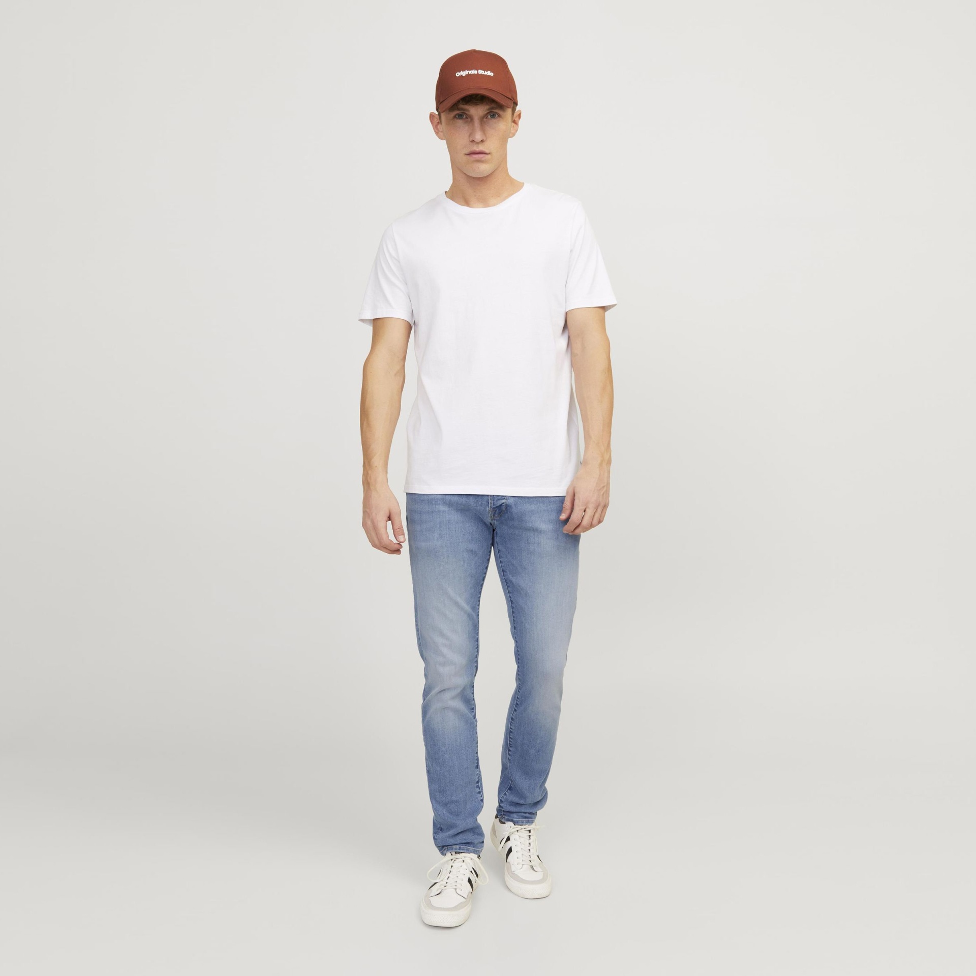 Homem com t-shirt branca, jeans azuis, sapatilhas brancas e boné castanho num fundo neutro.