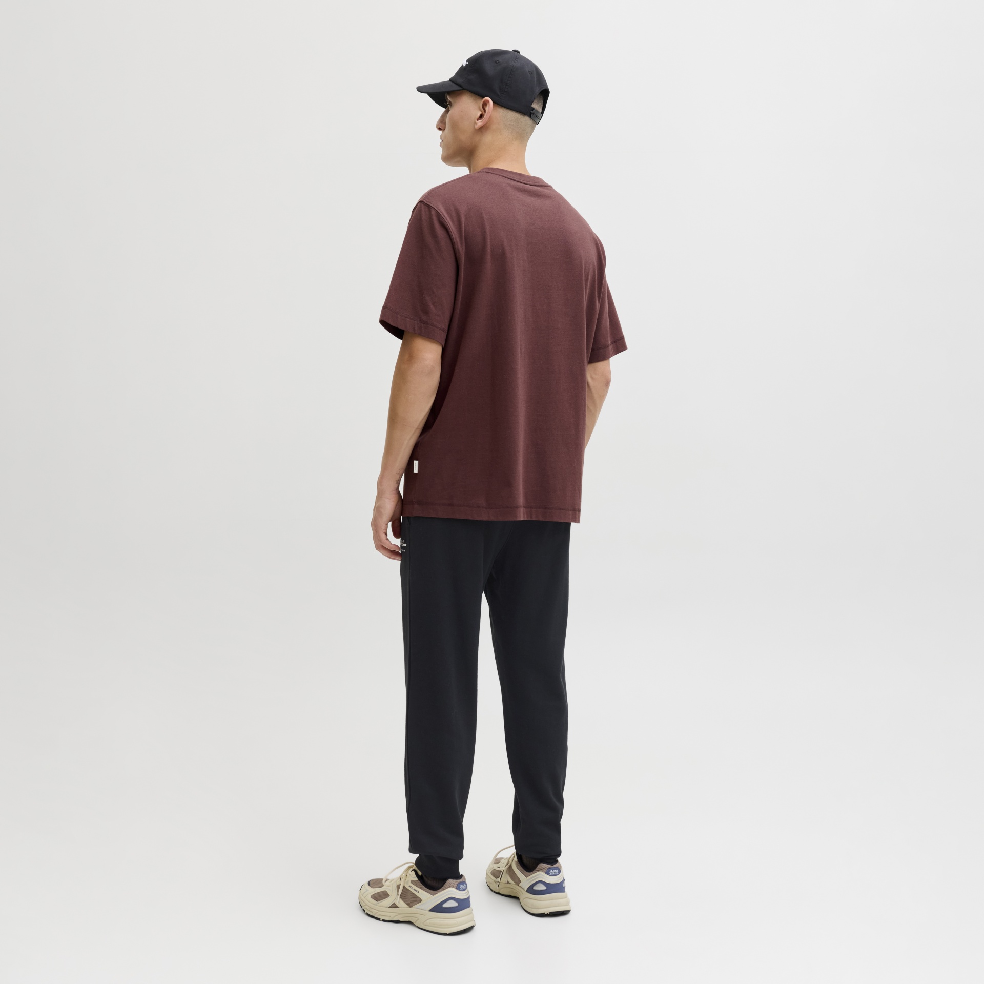 Pessoa de costas com t-shirt castanha, calças pretas, ténis bege com detalhes azuis e boné preto.