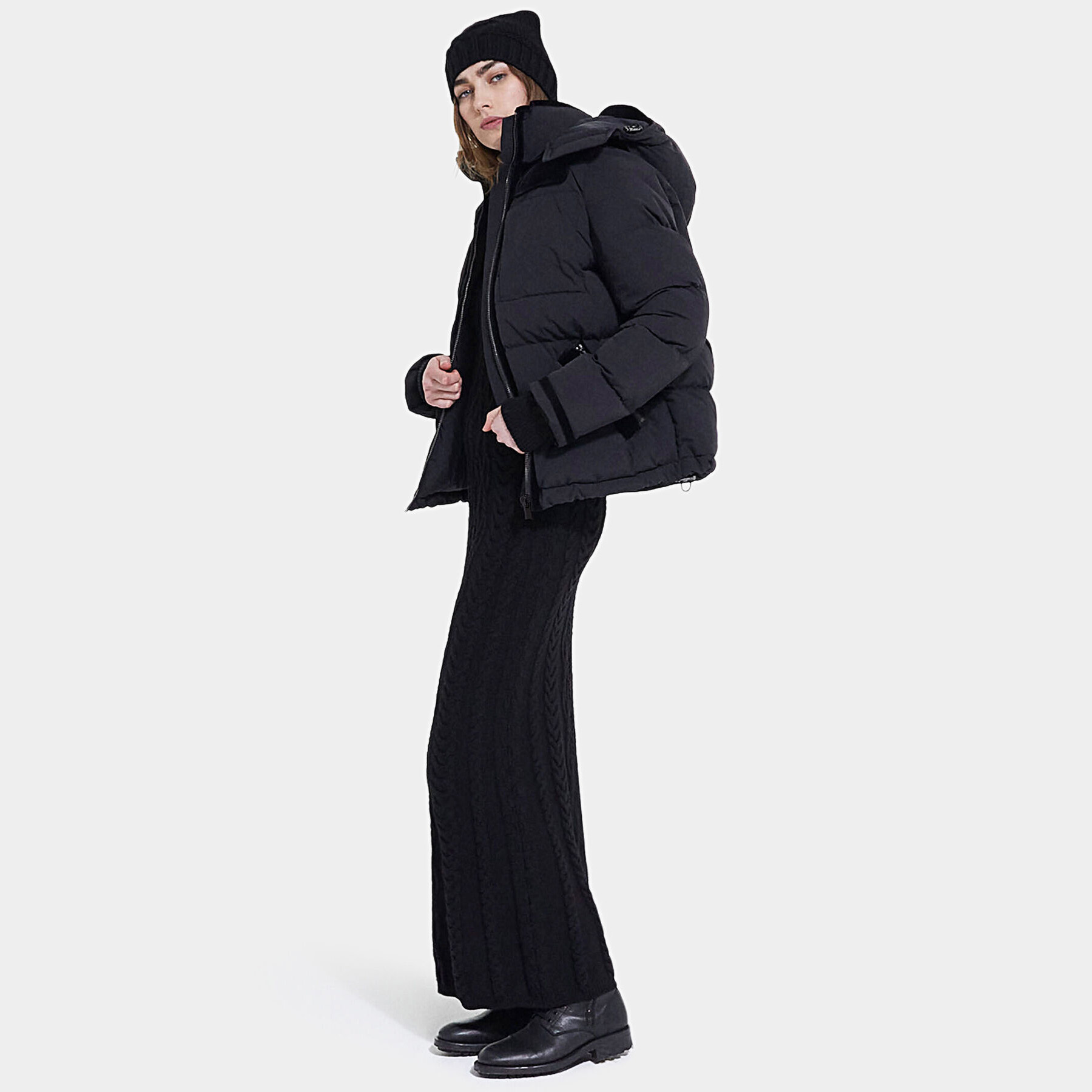 Mulher de casaco acolchoado preto, vestido longo de malha preta, bonnet preto e botas pretas.