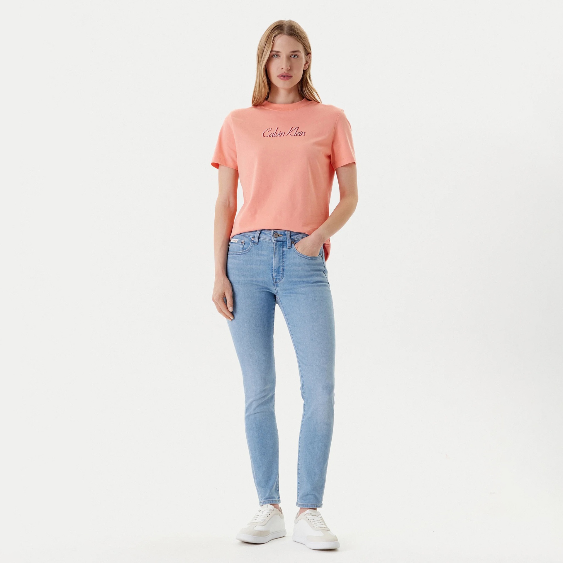 Mulher com t-shirt rosa Calvin Klein, jeans azuis claros e sapatos brancos em fundo branco