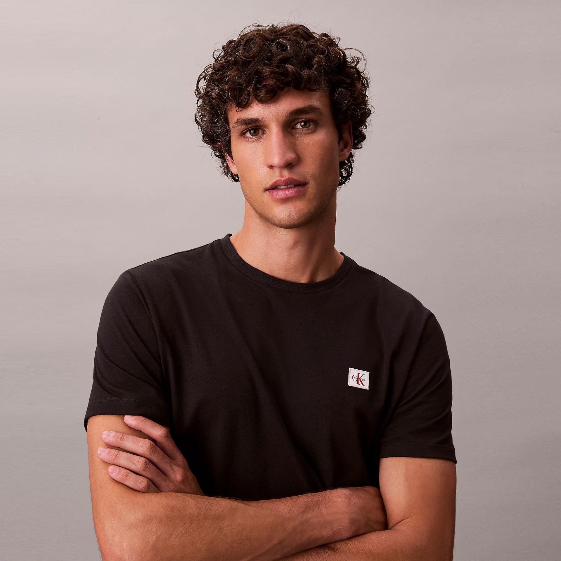 Homem com t-shirt preta de algodão com logo ck Calvin Klein no peito e braços cruzados.