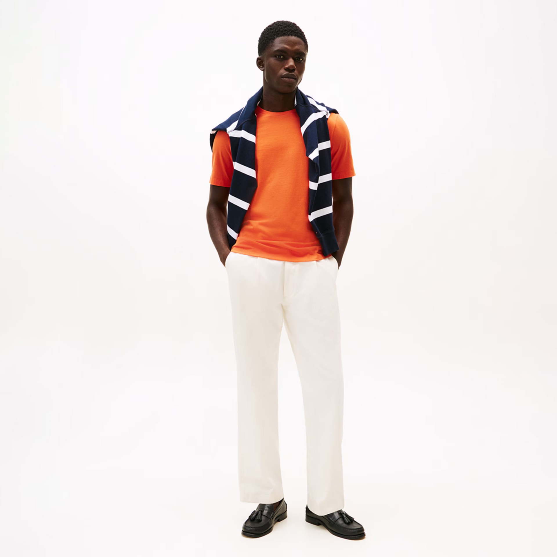 Homem vestido com t-shirt laranja, calças brancas, sapatos pretos e camisola azul com riscas brancas nos ombros.