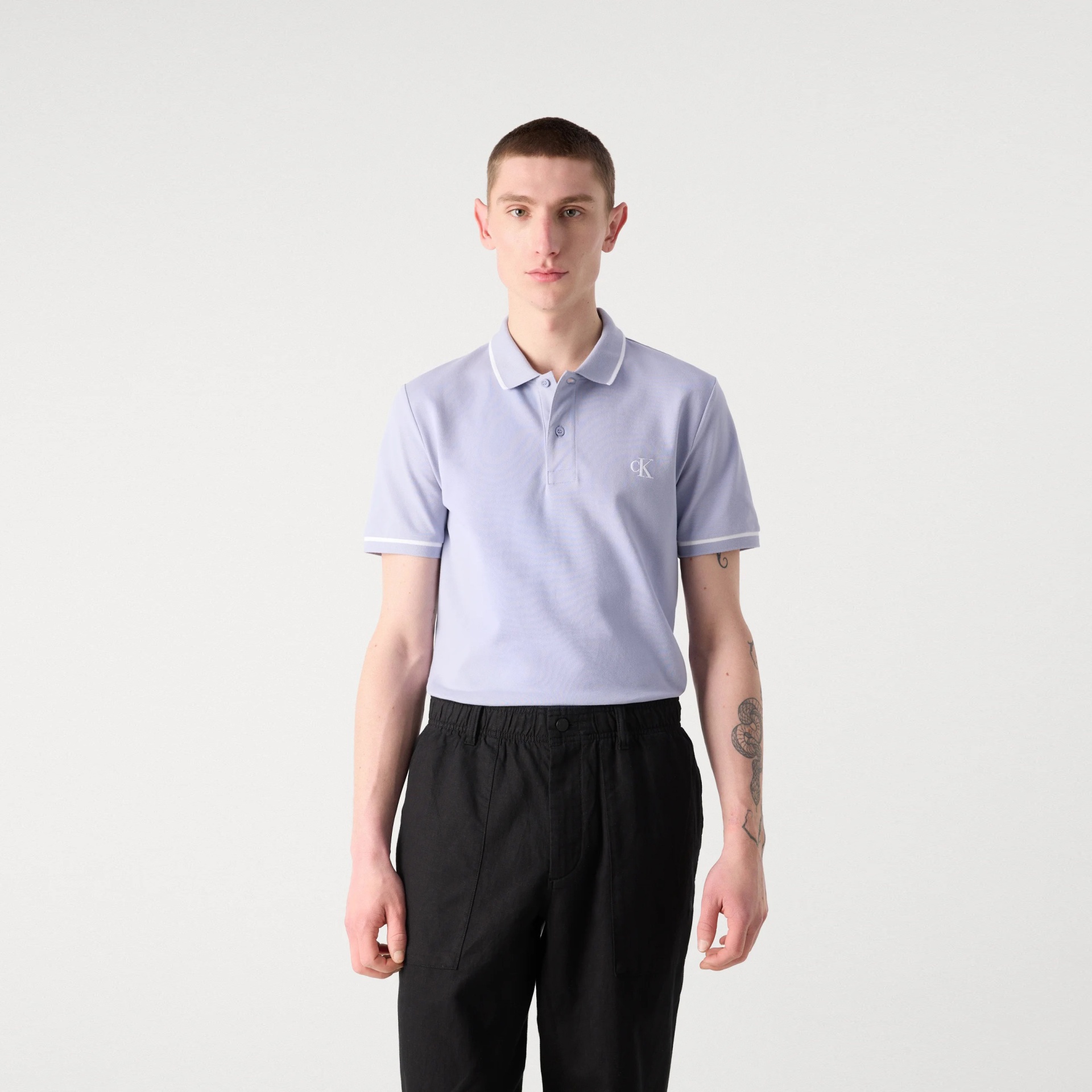 Homem com polo azul claro e calças pretas em fundo branco.