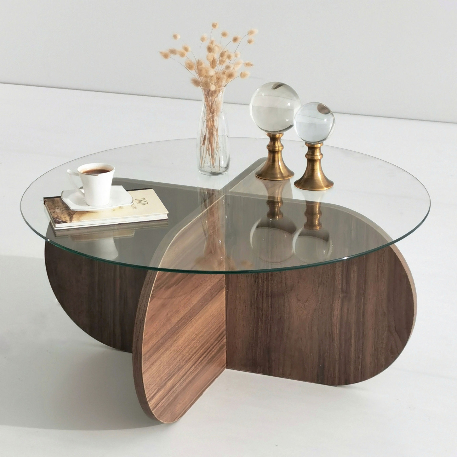Mesa de centro redonda com tampo de vidro e base de madeira, decorada com vaso, flores secas, esferas de vidro e chávena com livro