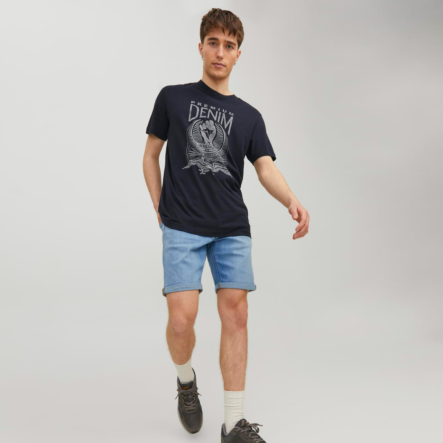 Homem com t-shirt preta estampada e calções azuis de ganga sobre fundo cinza claro