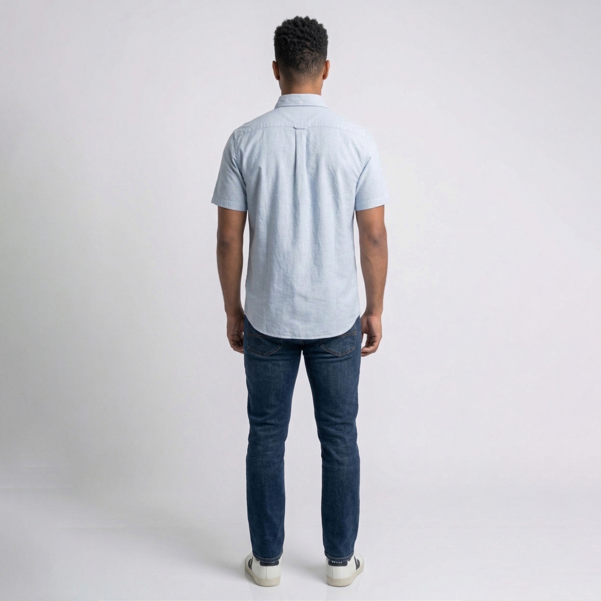 Homem de costas com camisa azul clara, jeans azul e ténis brancos em fundo branco