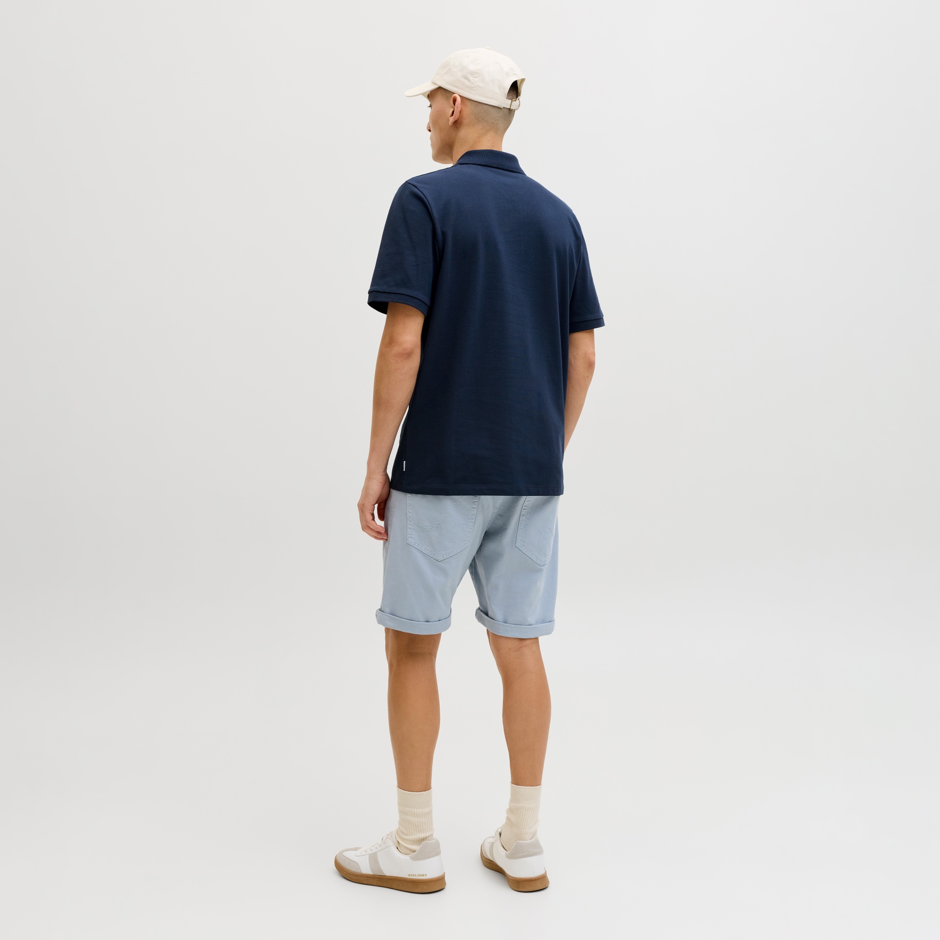 Homen com polo azul-marinho, calções azul claros, boné bege e ténis brancos sobre fundo branco