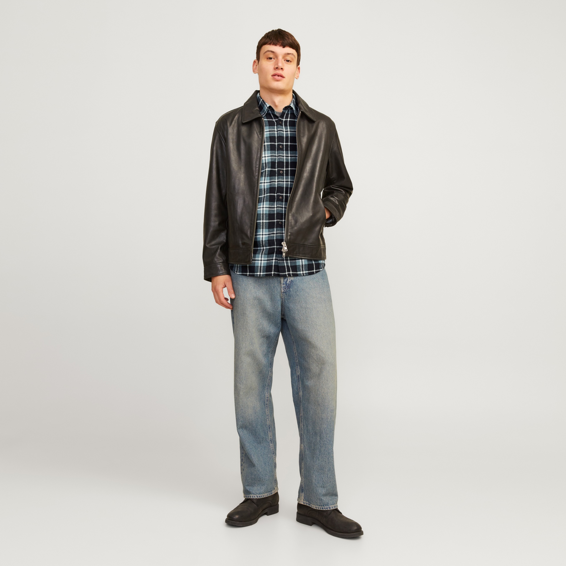 Homem com casaco de couro preto, camisa xadrez azul, jeans claros e botas pretas